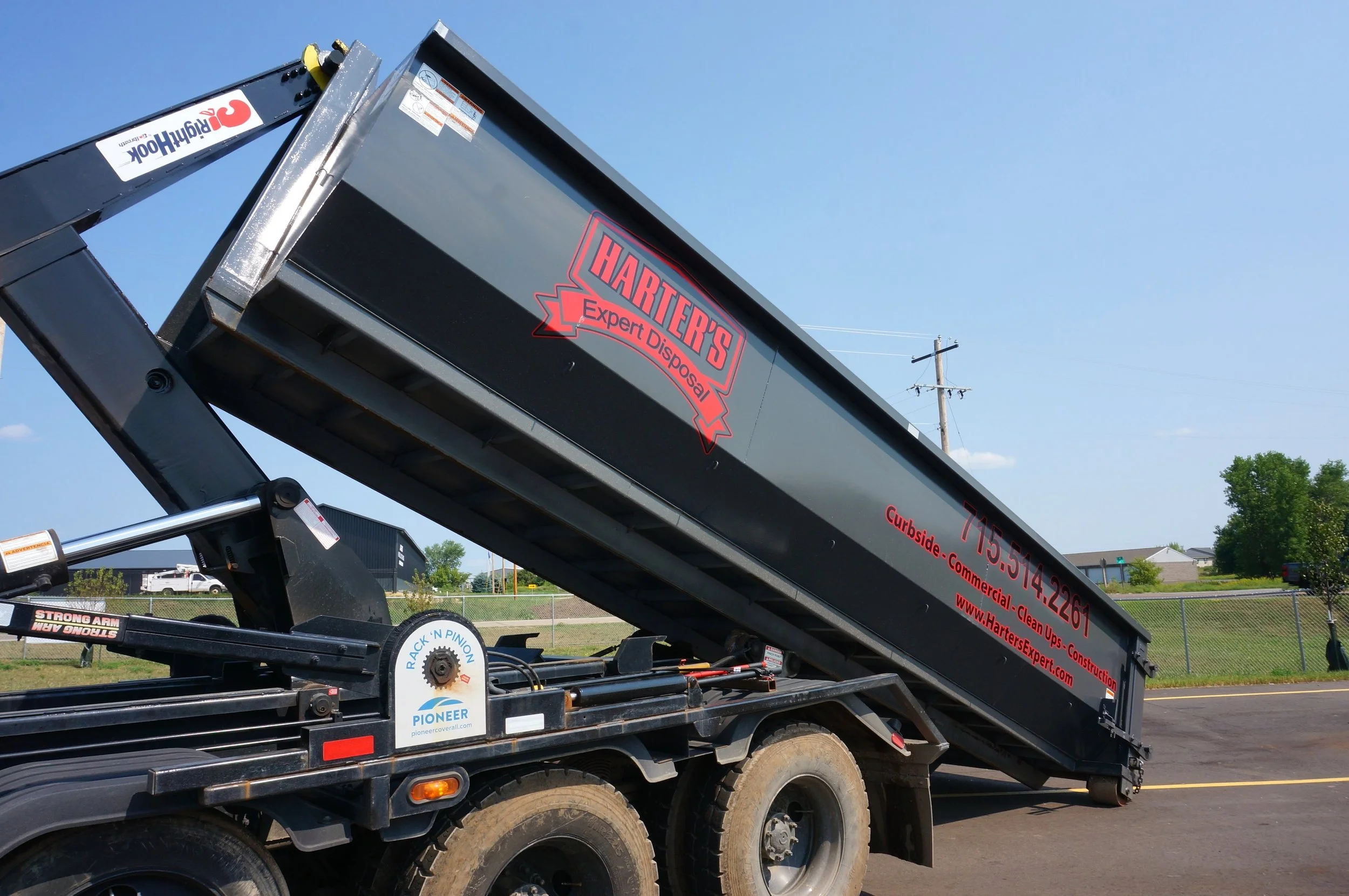A black flatbed truck with a hydraulic lift, branded with Hartzer's Expert Disposal, is parked in a lot on a clear day. The lift arm is extended upward, and the truck has various company stickers and contact information.