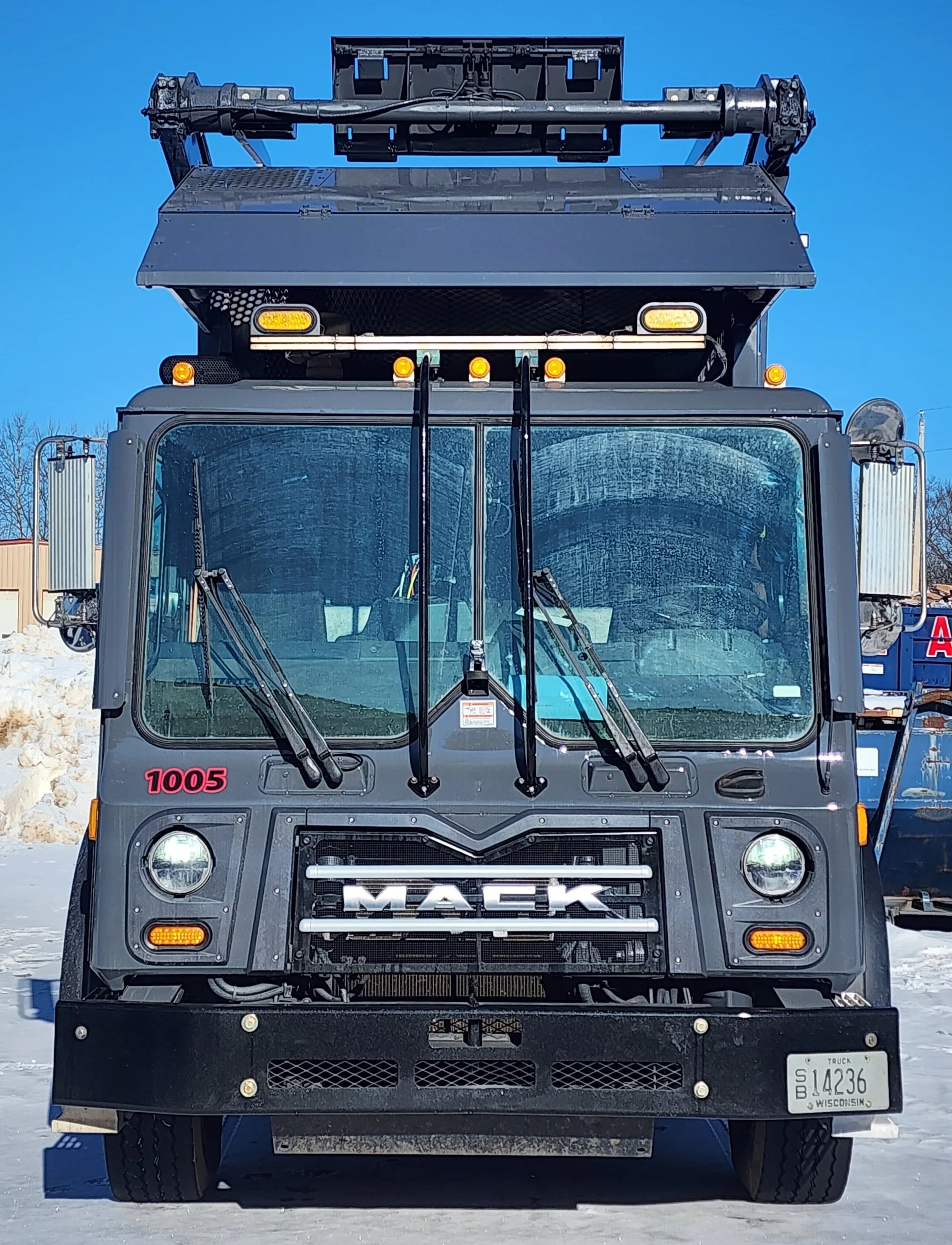Front view of a black Mack truck with chrome grille, headlights, and a Wisconsin license plate, parked outdoors under a clear blue sky.