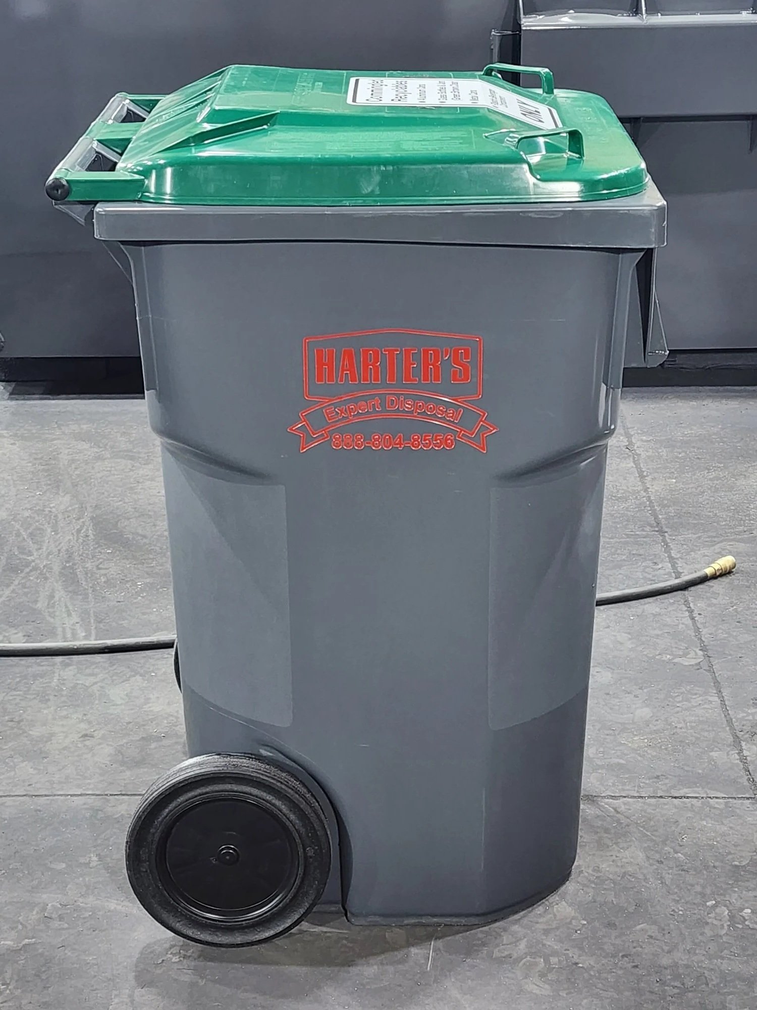 A gray trash bin with a green lid and black wheels, placed on a concrete floor, with a hose clip on the ground beside it. The bin has red and white lettering that reads 'Harter's Expert Disposal' and a phone number.