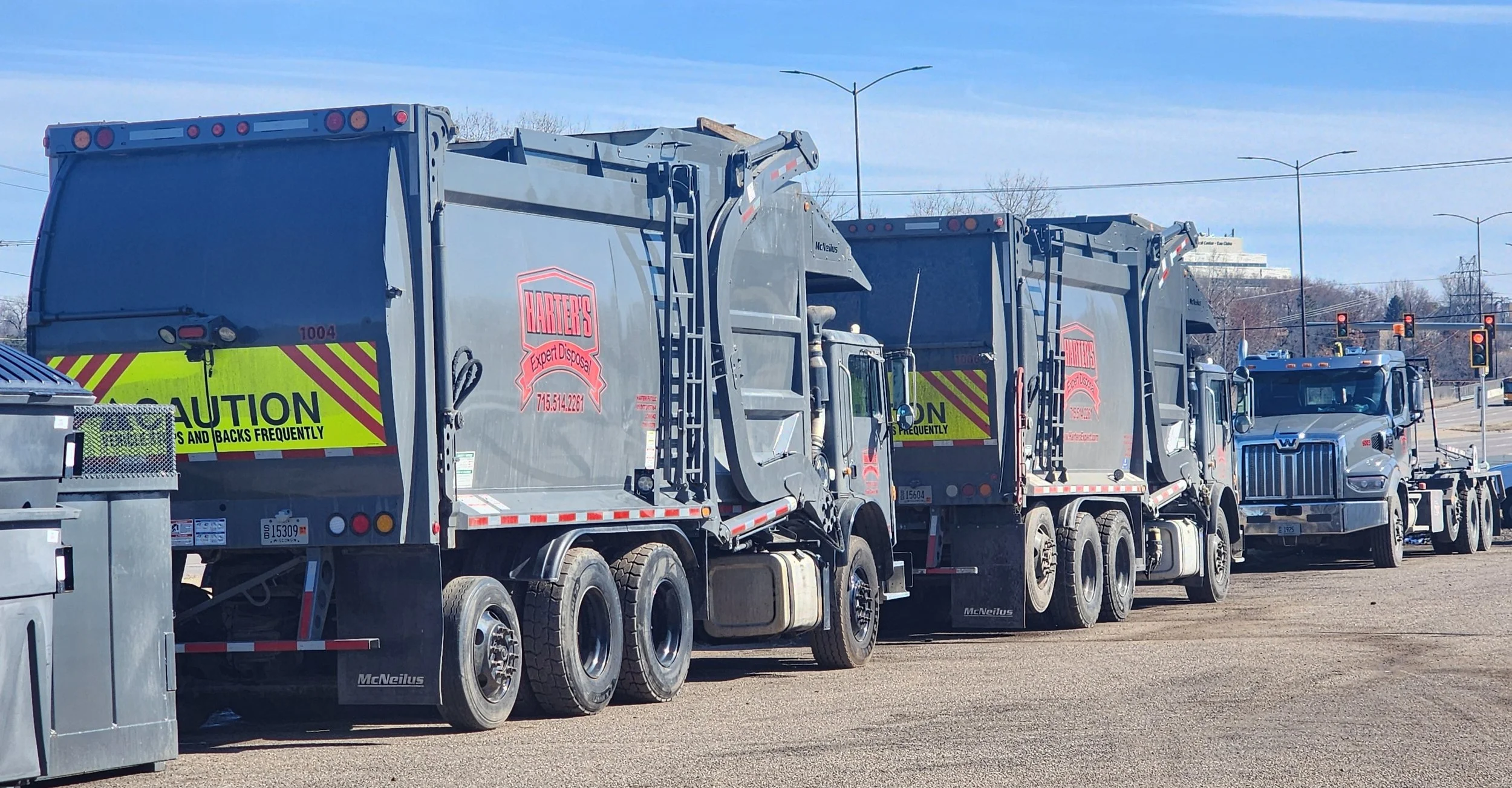Three waste collection trucks with gray bodies and yellow caution stripes parked in a lot, with a pickup truck behind them and power lines and trees in the background.
