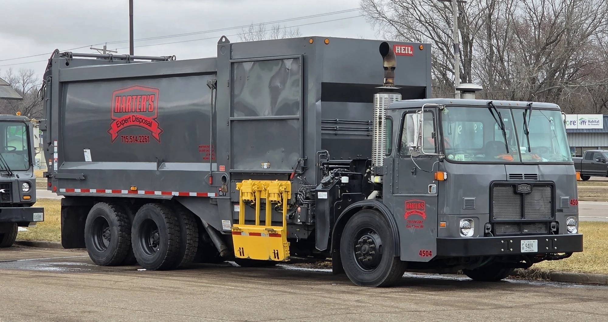 Gray waste collection truck with red and black company logo and contact information parked on a street with trees and buildings in the background.