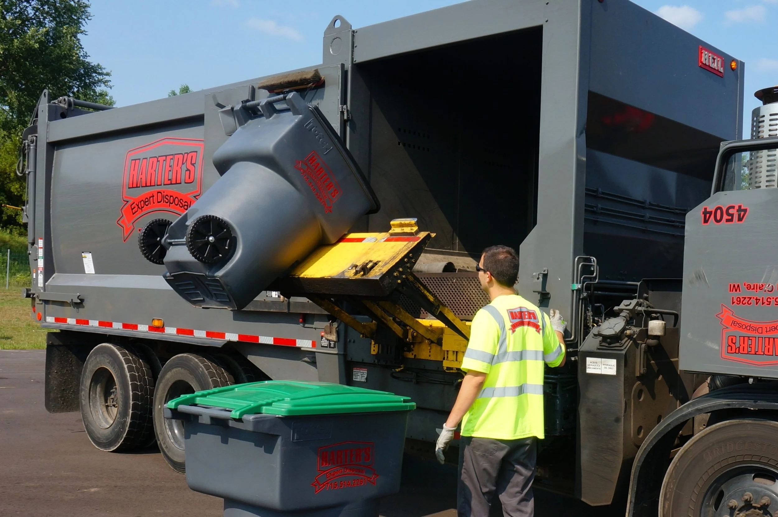 A large gray trash disposal truck with the logo 'Harter's Expert Disposal' on the side, with a person in a bright yellow safety vest walking nearby. The truck has a waste bin in the process of being emptied into the container, and there are green trash cans at the bottom of the image.