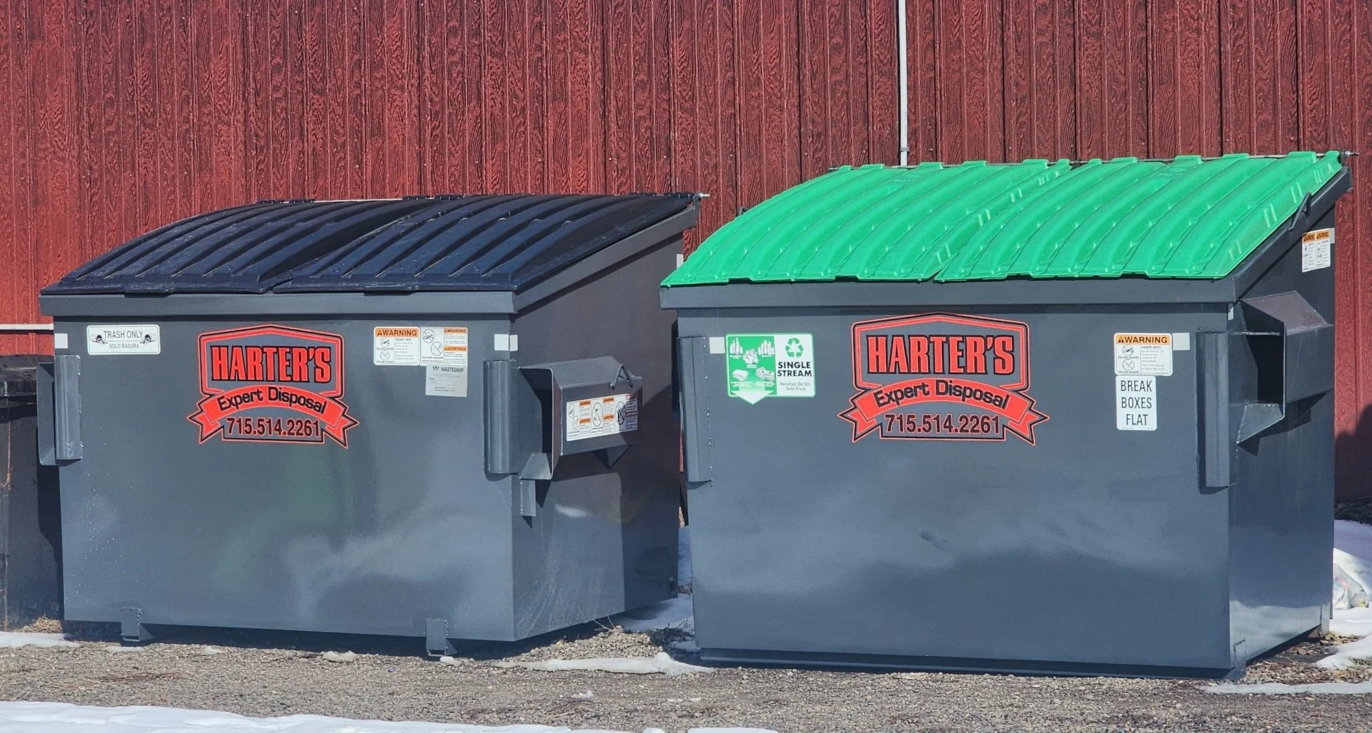 Two large trash dumpsters with black and green lids placed against a red wooden wall. The black lid dumpster has a white label that reads 'TRASH ONLY' and a company logo, while the green lid dumpster has a recycling symbol and related labels.