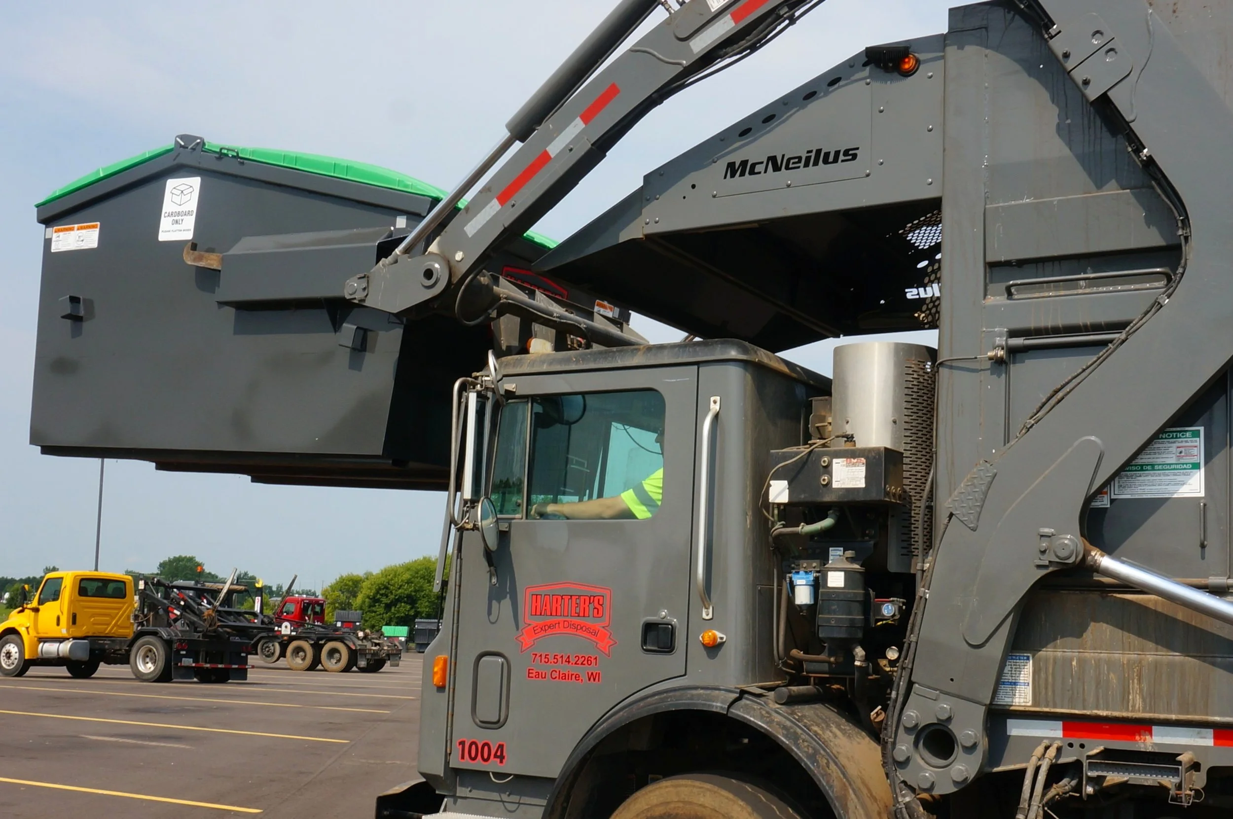 A gray trash truck with green and black accents is parked in a lot alongside other trucks. The truck has the company name 'Harter's' on the side and contact information for Eau Claire, Wisconsin. In the background, other trucks and moving equipment are visible under a cloudy sky.