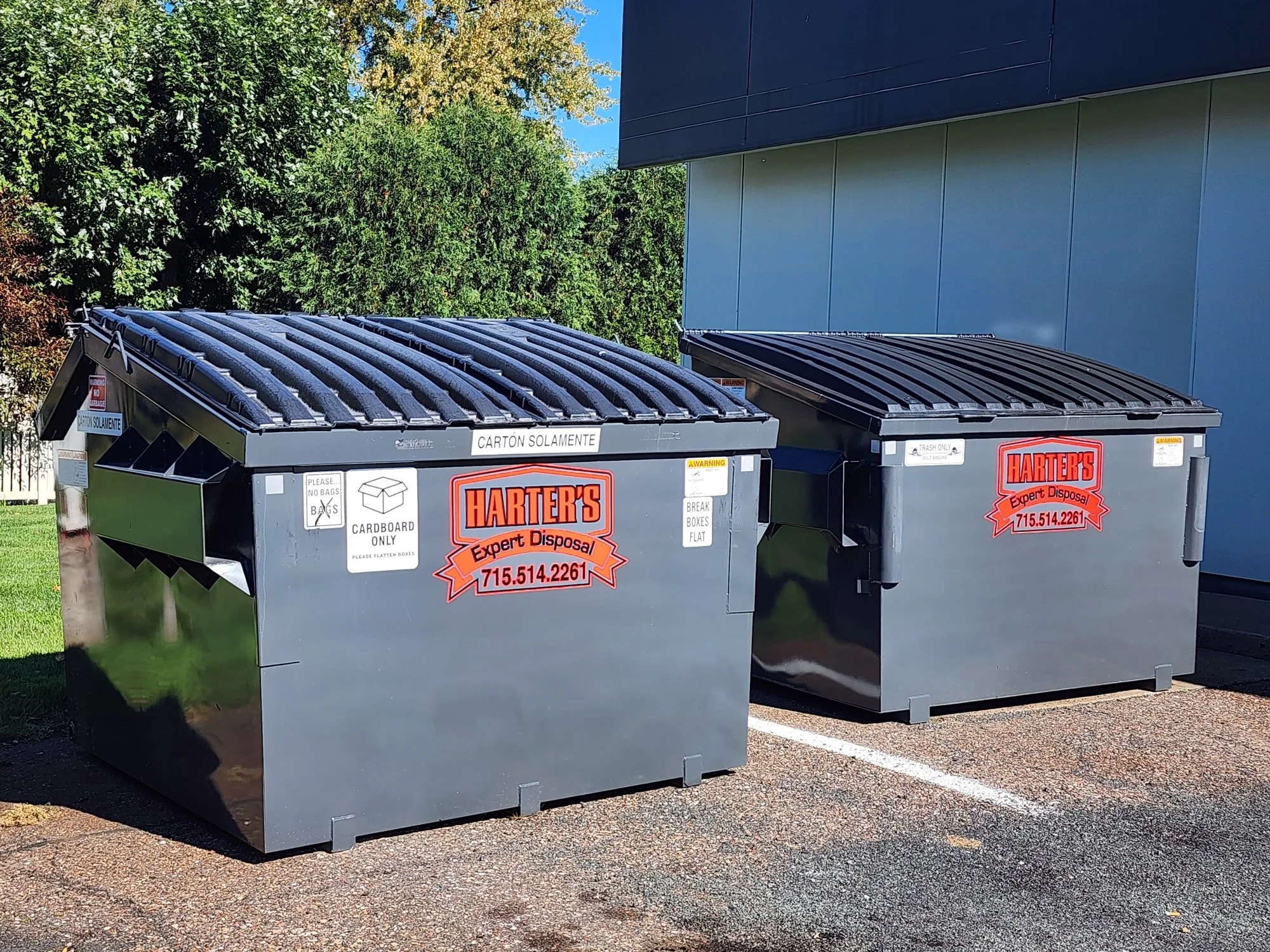 Two gray trash disposal bins with black lids outside, labeled for cardboard and trash only, placed side by side on a parking lot with green trees and a building in the background.