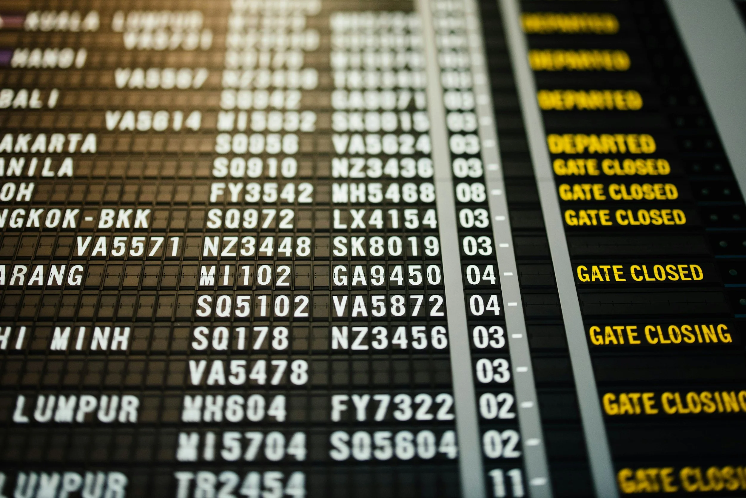 An airport departure and arrival flight information display with flight numbers, destinations, and status updates including "DEPARTED" and "GATE CLOSED" in yellow text on black background.