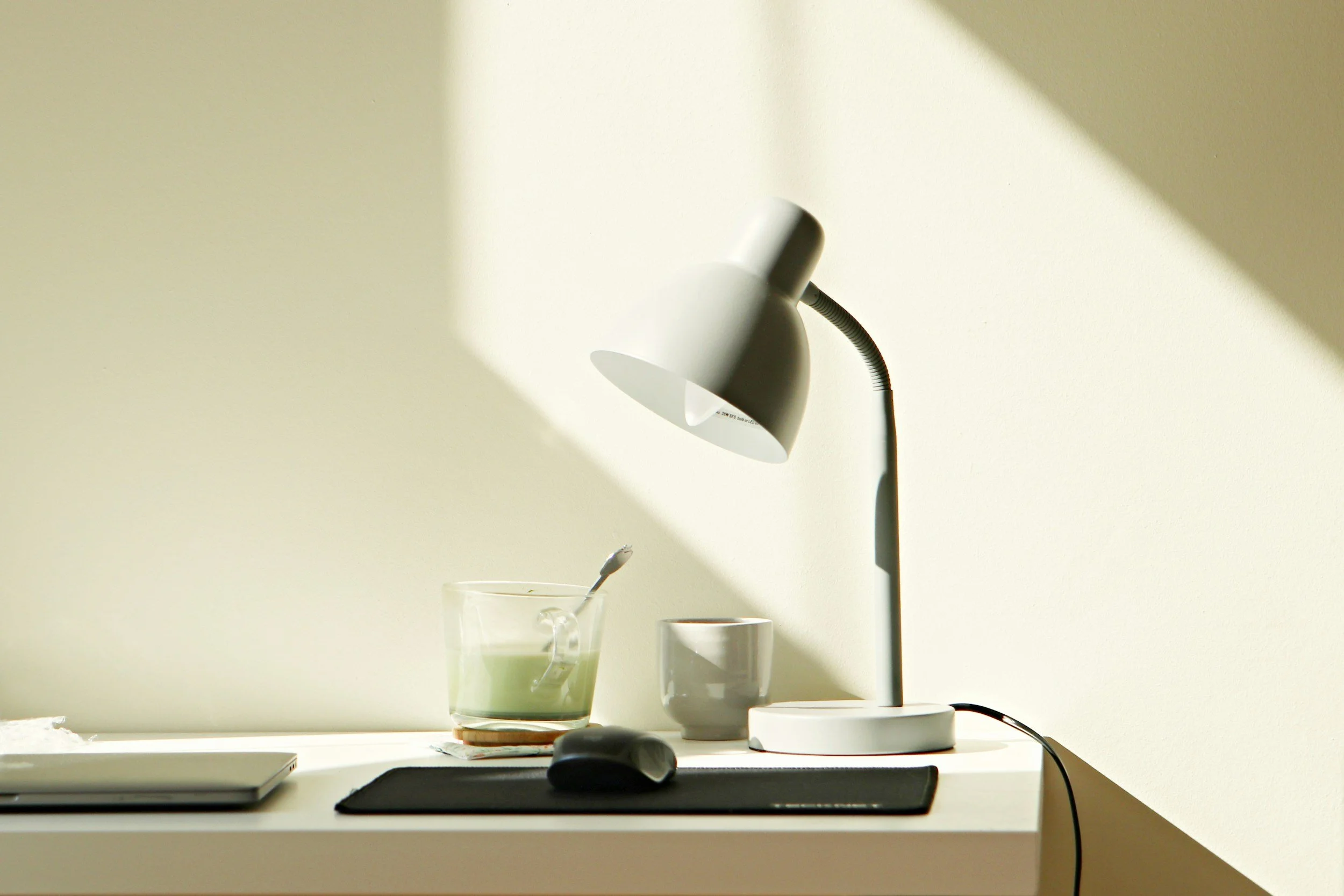 A minimalist desk setup with a modern white desk lamp, a glass of green tea with a spoon, an empty white cup, a closed laptop, a computer mouse, and a black mouse pad, with sunlight casting shadows on a cream-colored wall.