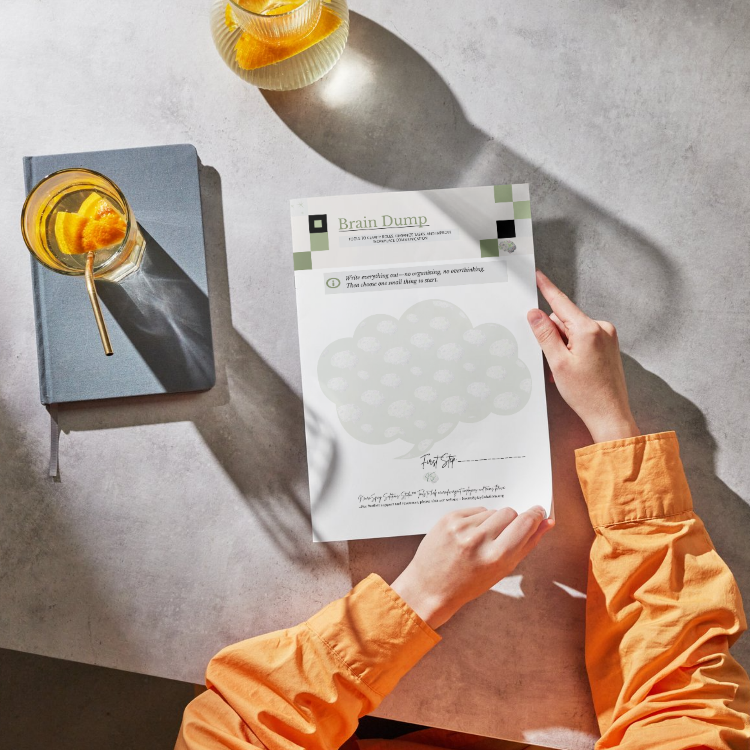 A person with orange sleeves holding a worksheet titled "Brain Dump" on a light-colored table. There is a book and a glass with a drink and lemon peel beside it. The worksheet has a cloud shape for writing.