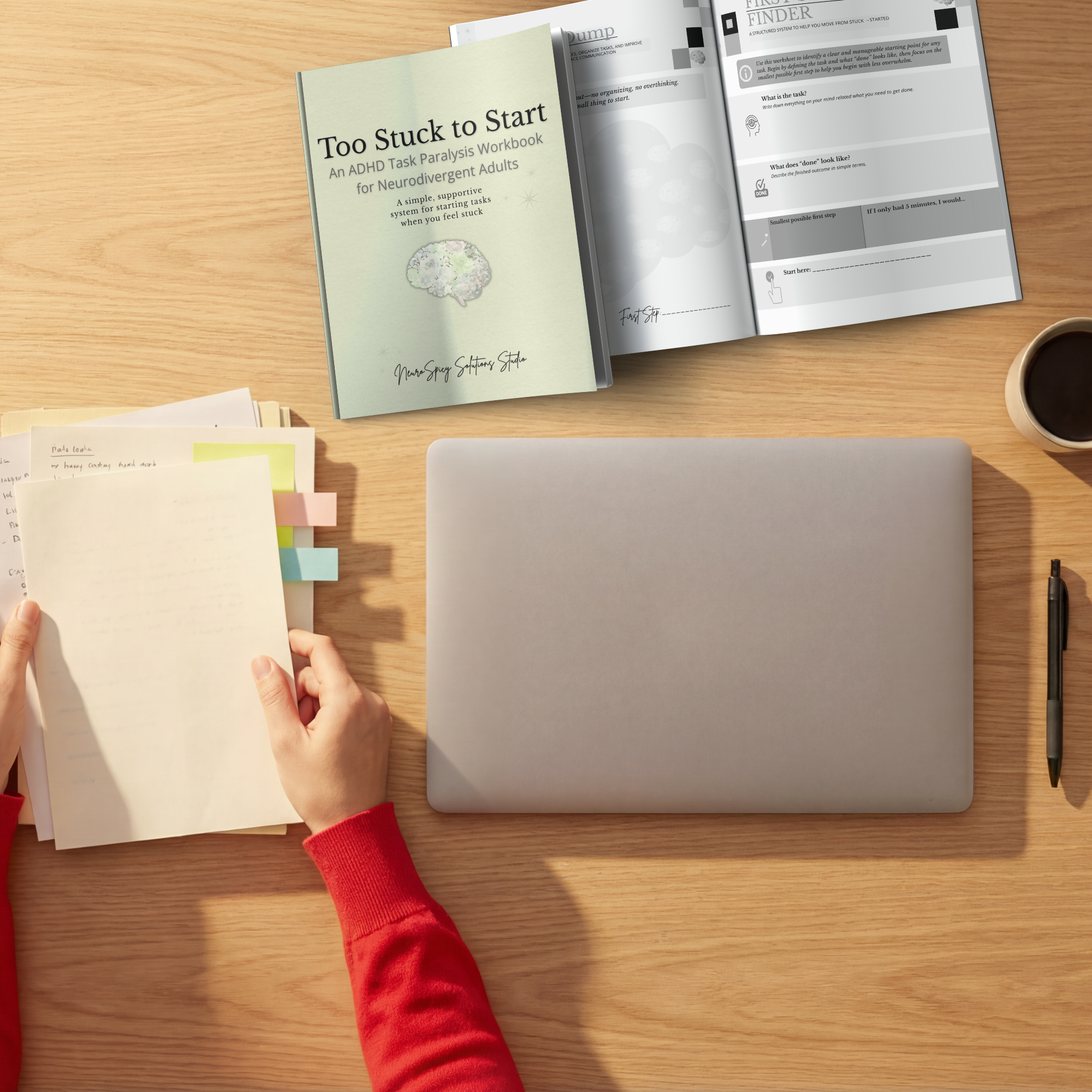 Open workbook titled 'Too Stuck to Start' on a wooden desk with a hand holding a small notepad, a closed silver laptop, a black pen, and several sticky notes in front of the hand. The workbook appears to be about helping neurodivergent adults start tasks.