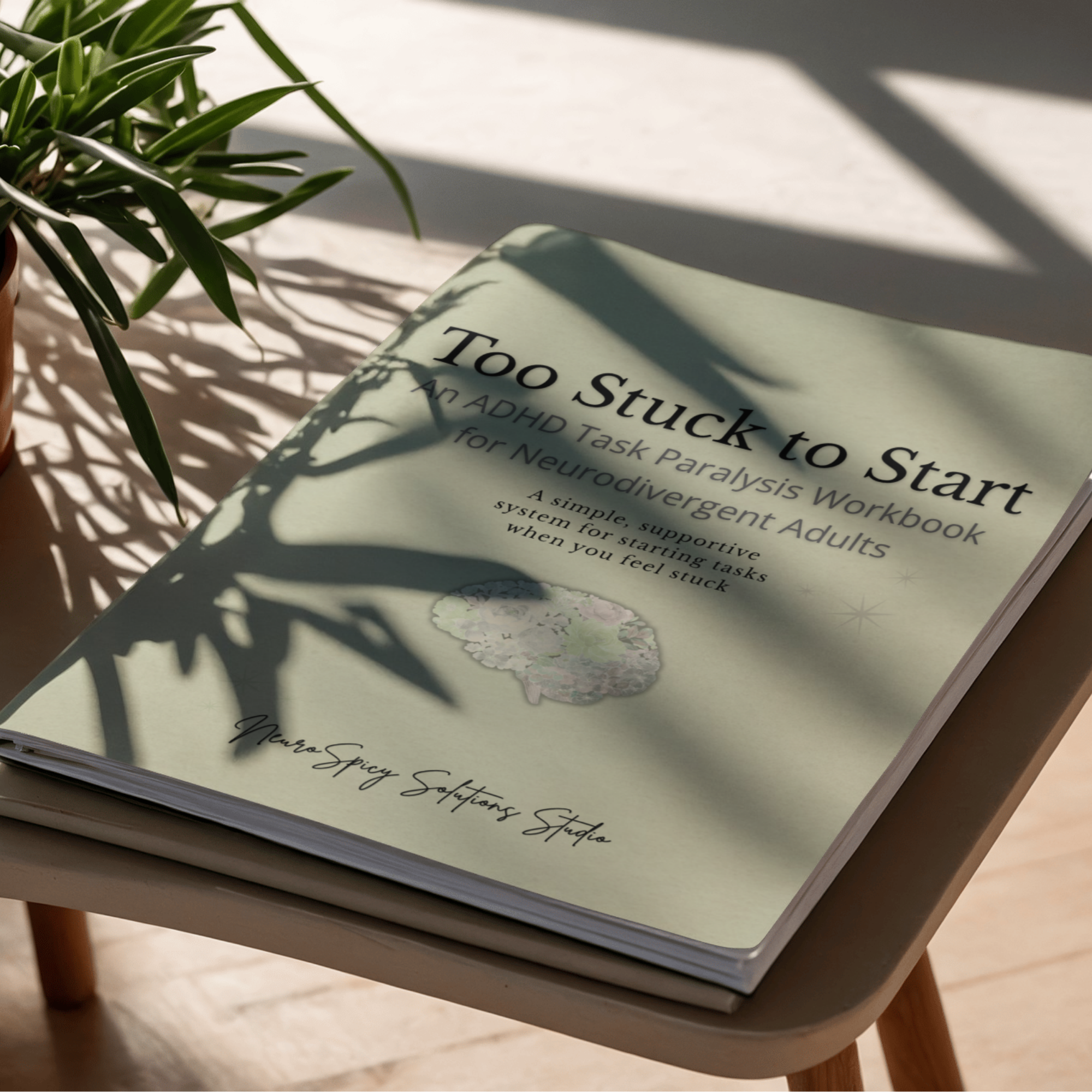 Close-up of a potted plant with long, green leaves on a wooden table next to a book titled 'Too Stuck to Start' in shadowed light.