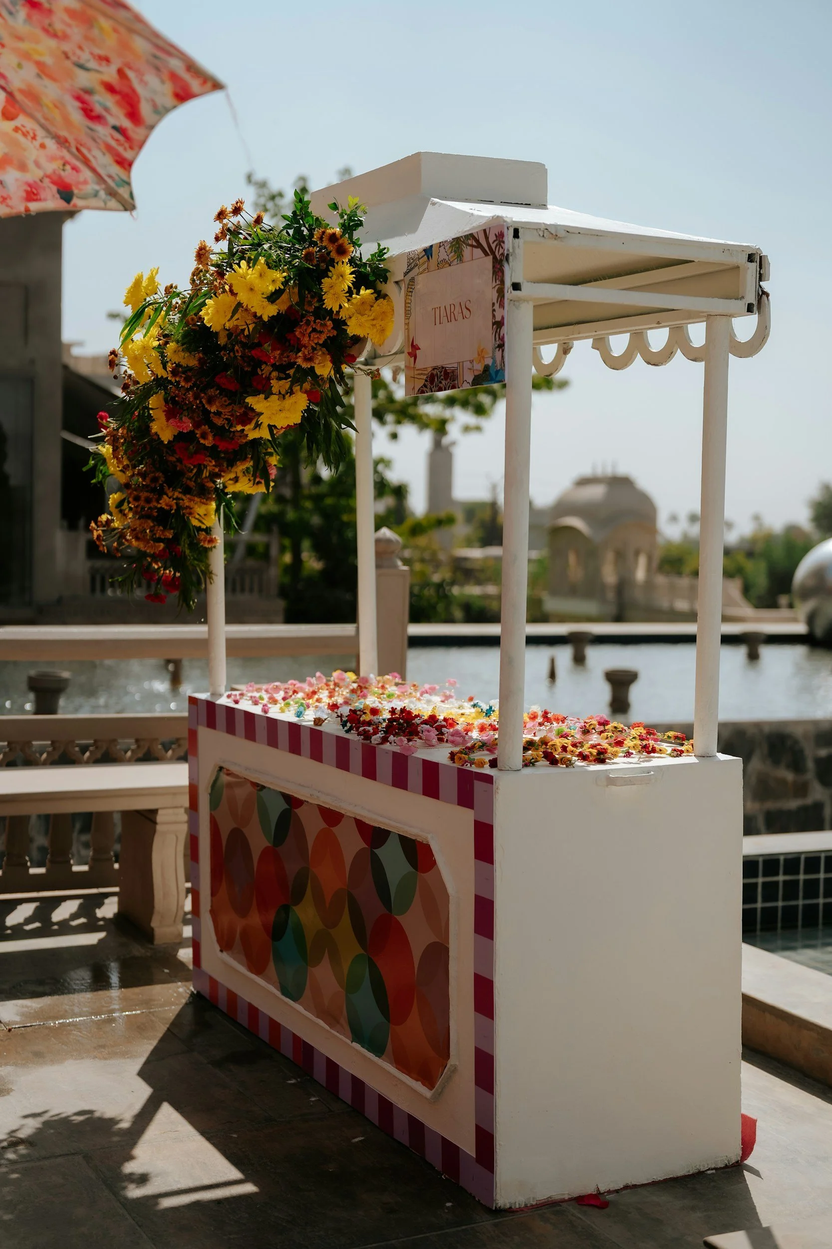 A small, colorful street cart decorated with a sign labeled 'Tiaras' and adorned with a large bouquet of yellow and orange flowers, situated outdoors near a water body with historic buildings in the background.