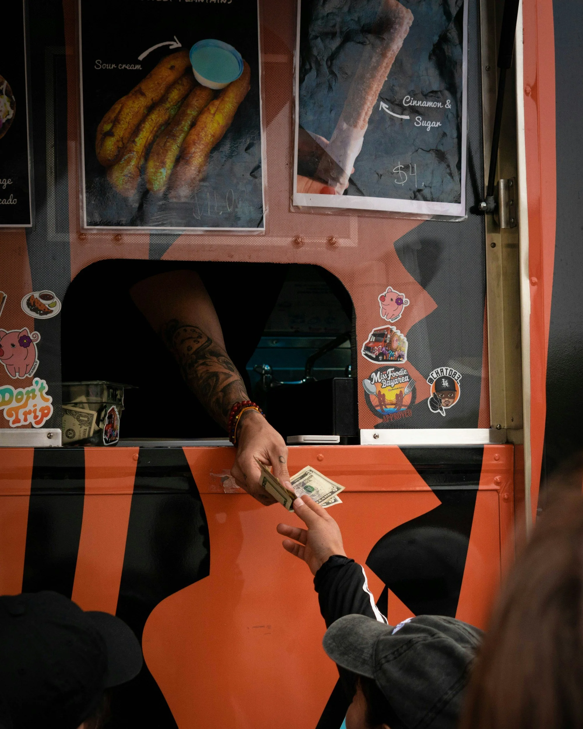 A person is handing money to a cashier at a food truck with stickers on the front. The food truck has images and prices of churros, one with sour cream and the other with cinnamon and sugar.