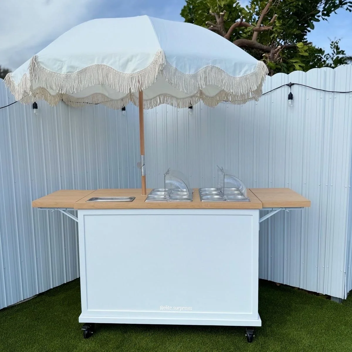 Outdoor ice cream or food cart with a large white umbrella and a wooden counter, set against a white fence and green grass, with tree branches overhead.