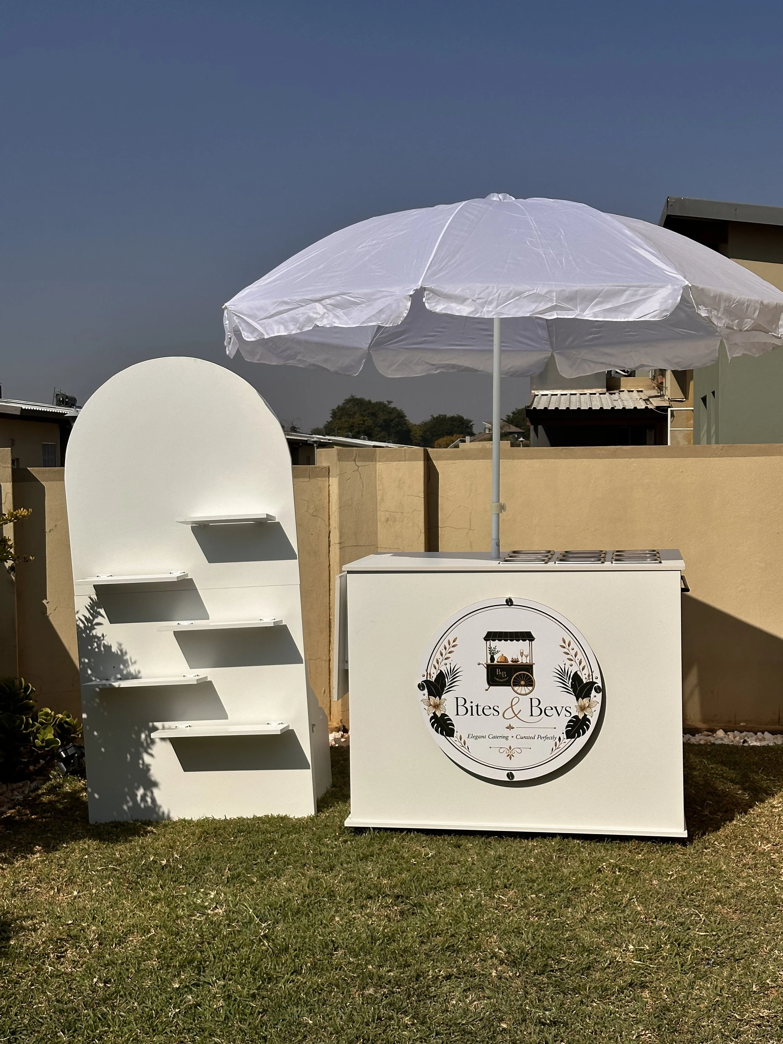 Outdoor setup for catering or event service featuring a white umbrella, a chafing dish on a counter with a sign reading "Bites & Bevs," and a white ladder-style shelf against a beige wall.
