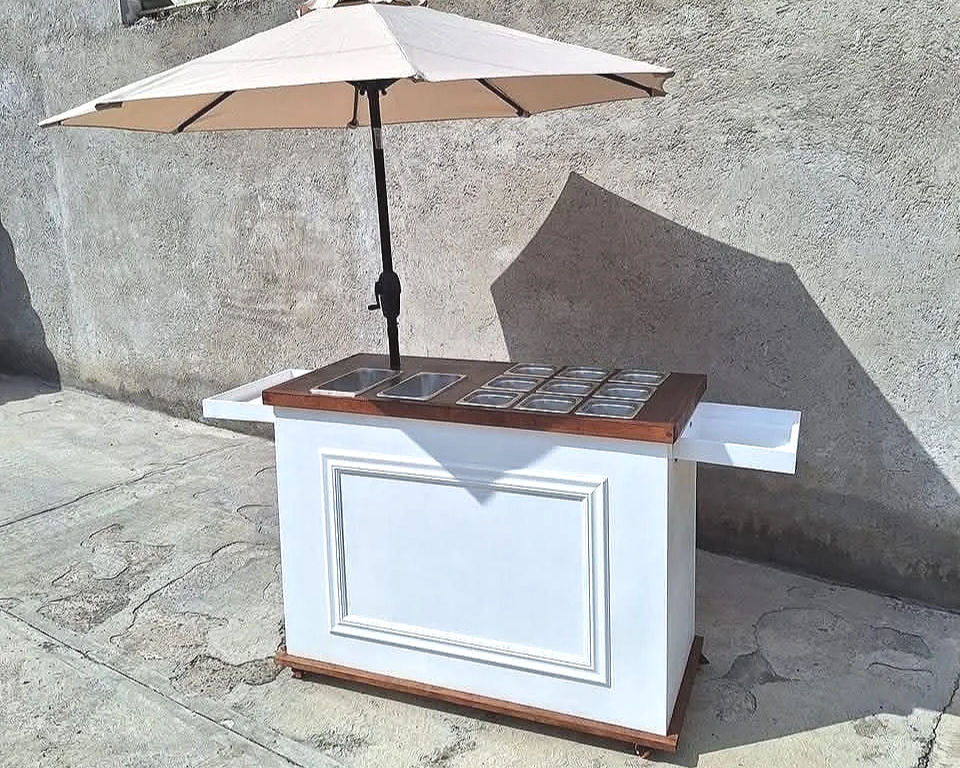 A small outdoor food cart with a beige umbrella, two stainless steel compartments, and a wooden countertop, positioned against a concrete wall on a sidewalk.