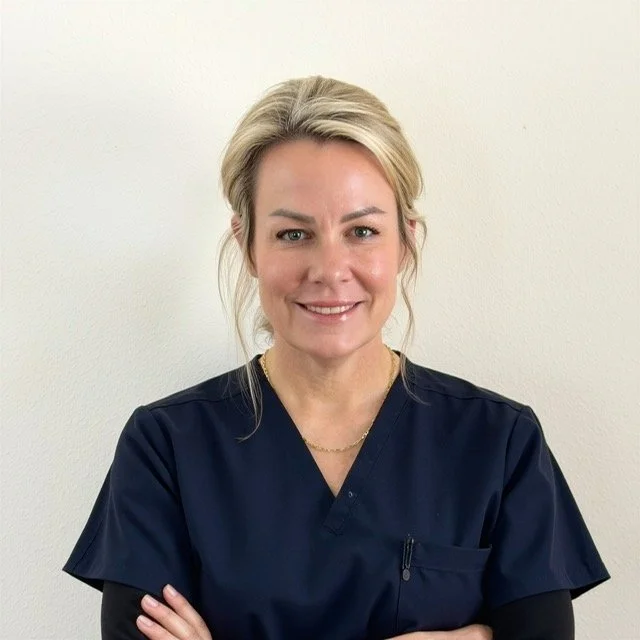 A woman with blonde hair tied back, wearing navy blue medical scrubs and a gold necklace, smiling and standing against a plain white wall. Solara Med Aesthetics in Oceanside, CA offers Botox, HA fillers, microneedling, PRP/PRF, biostimulators, and IV