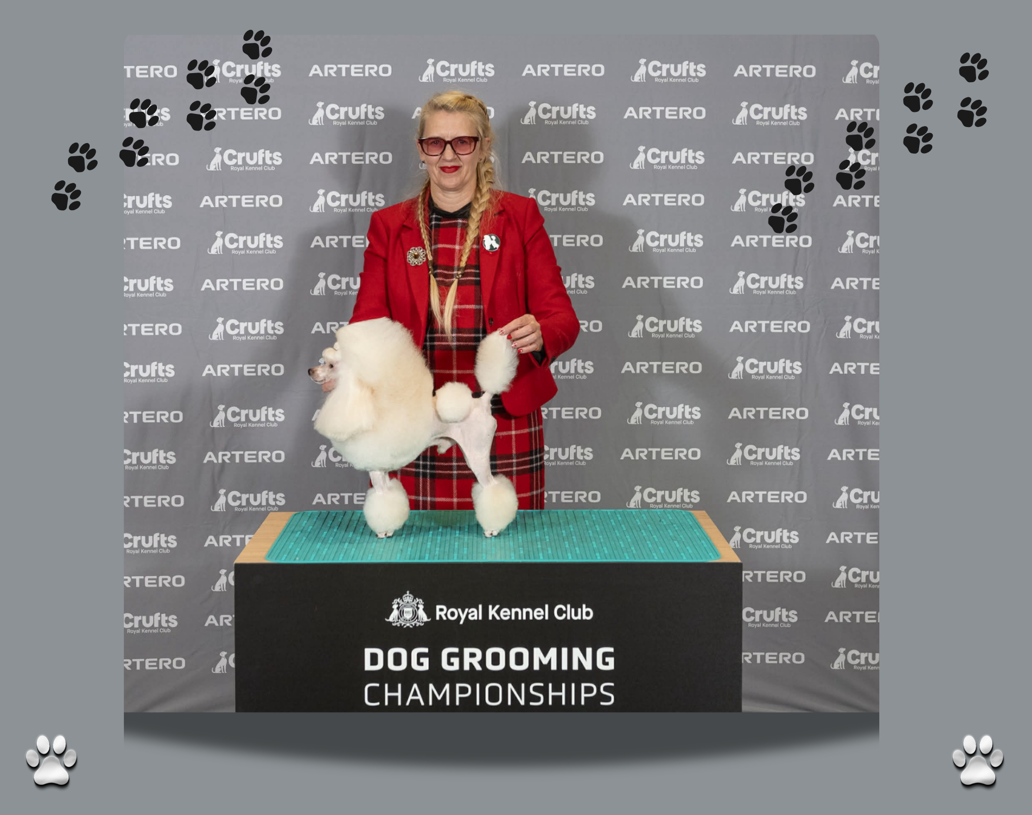 At a dog grooming championship event, a woman in red plaid and red blazer with sunglasses and blonde braided hair stands at a grooming table with a white poodle. The background features a grey banner with logos of Crufts Royal Kennel Club, Artero, and Tero, and black paw print decorations.