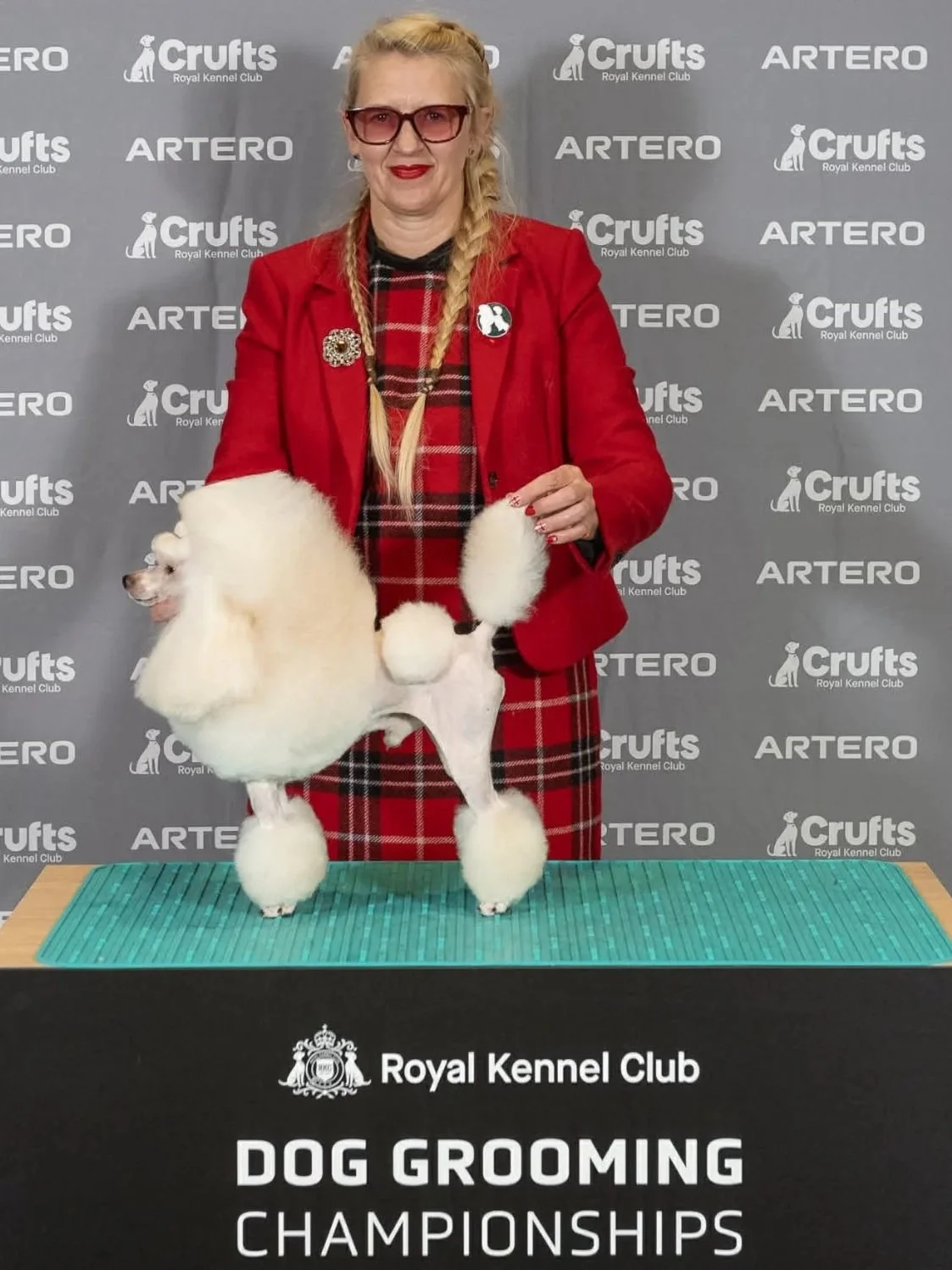 A woman with blonde hair in braids, wearing glasses, a red blazer, and a red plaid dress, stands behind a grooming table with a groomed white poodle. The background shows a gray step-and-repeat banner with logos, including 'Crufts', 'Artero', and 'Royal Kennel Club'. The table has a black sign reading 'Royal Kennel Club Dog Grooming Championships'.