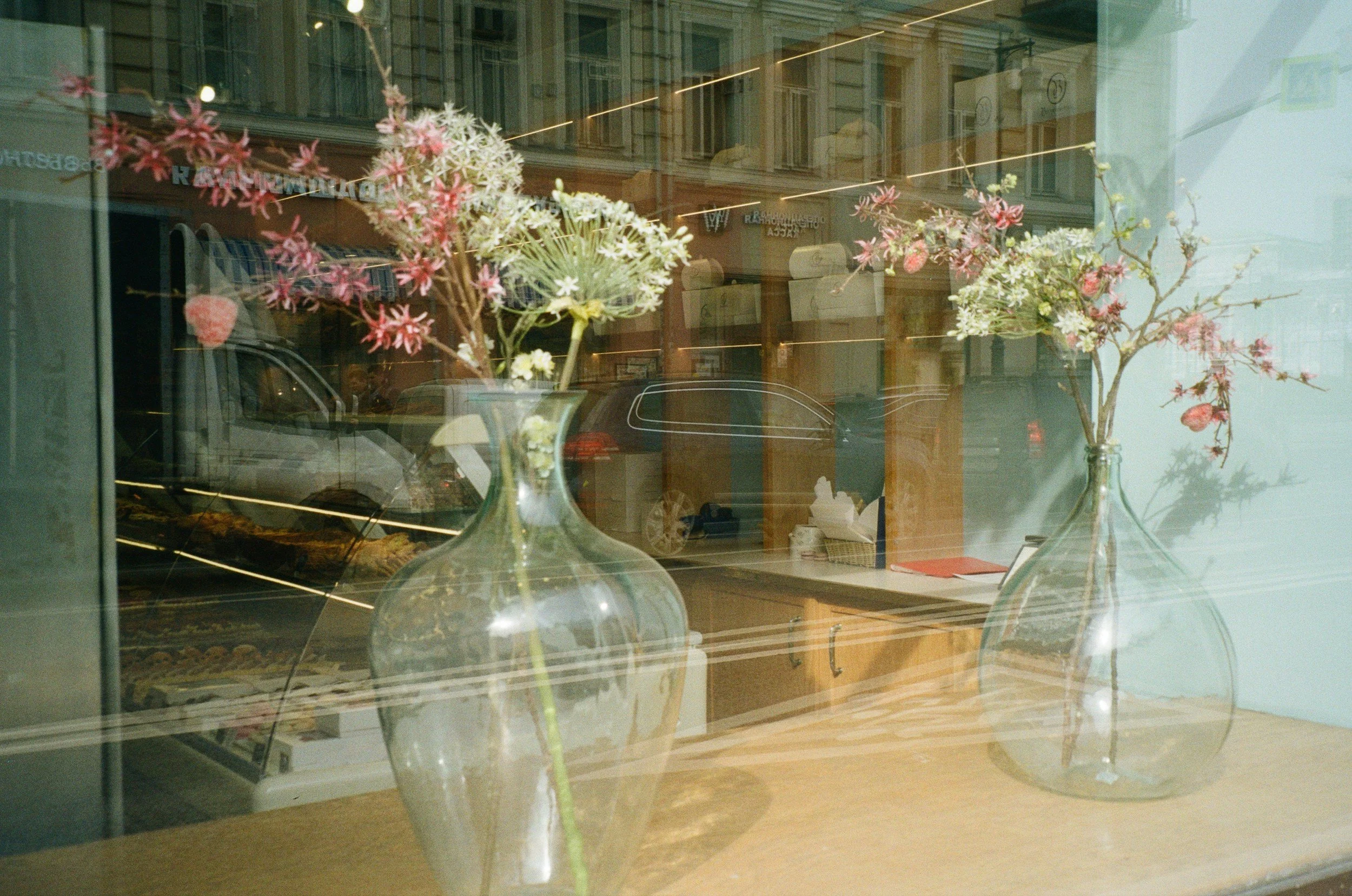 Two glass vases with pink and white flowers placed on a wooden surface, viewed through a window with reflections of the street and buildings outside.
