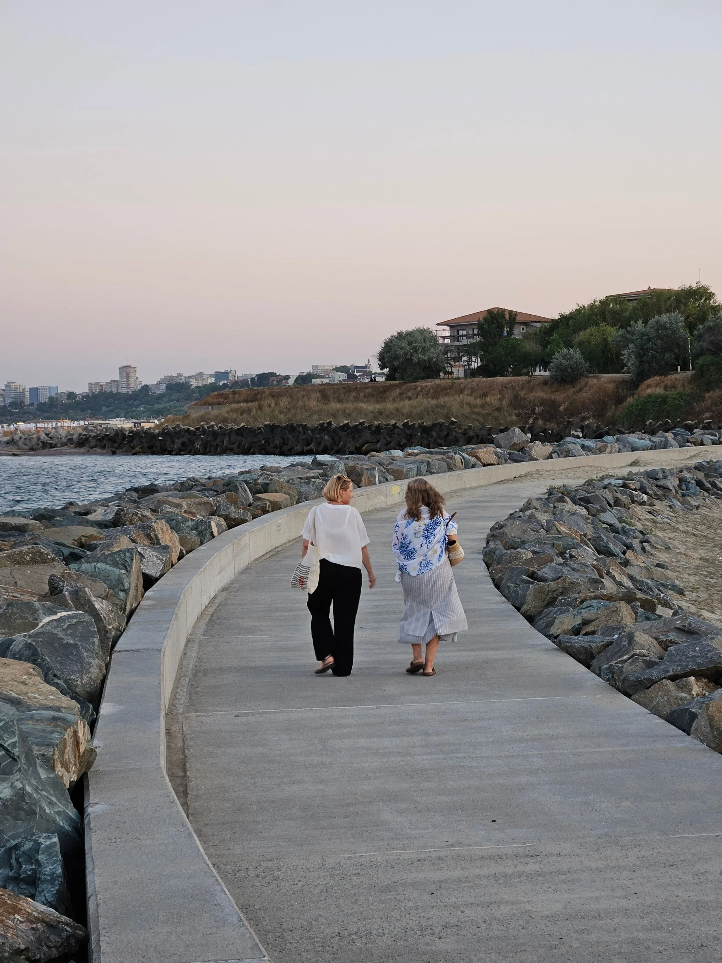 Two women walking along a curved concrete pathway beside a rocky shoreline at dusk, with a cityscape and houses on a hill in the background.