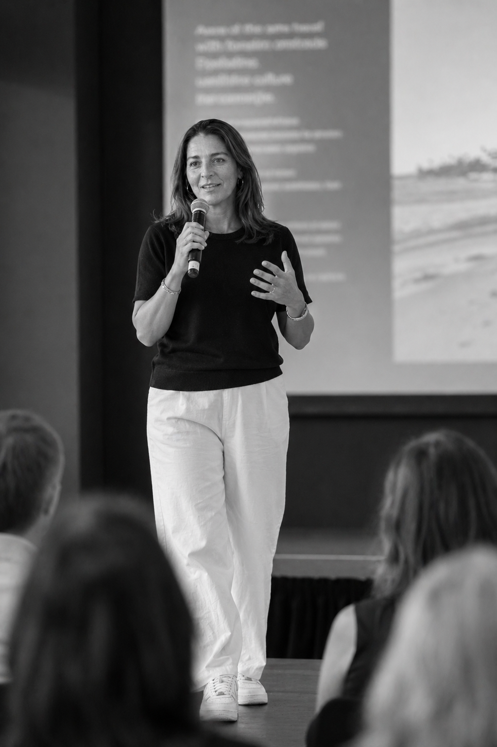 Black and white photo of a woman holding a microphone and speaking to an audience during a presentation.
