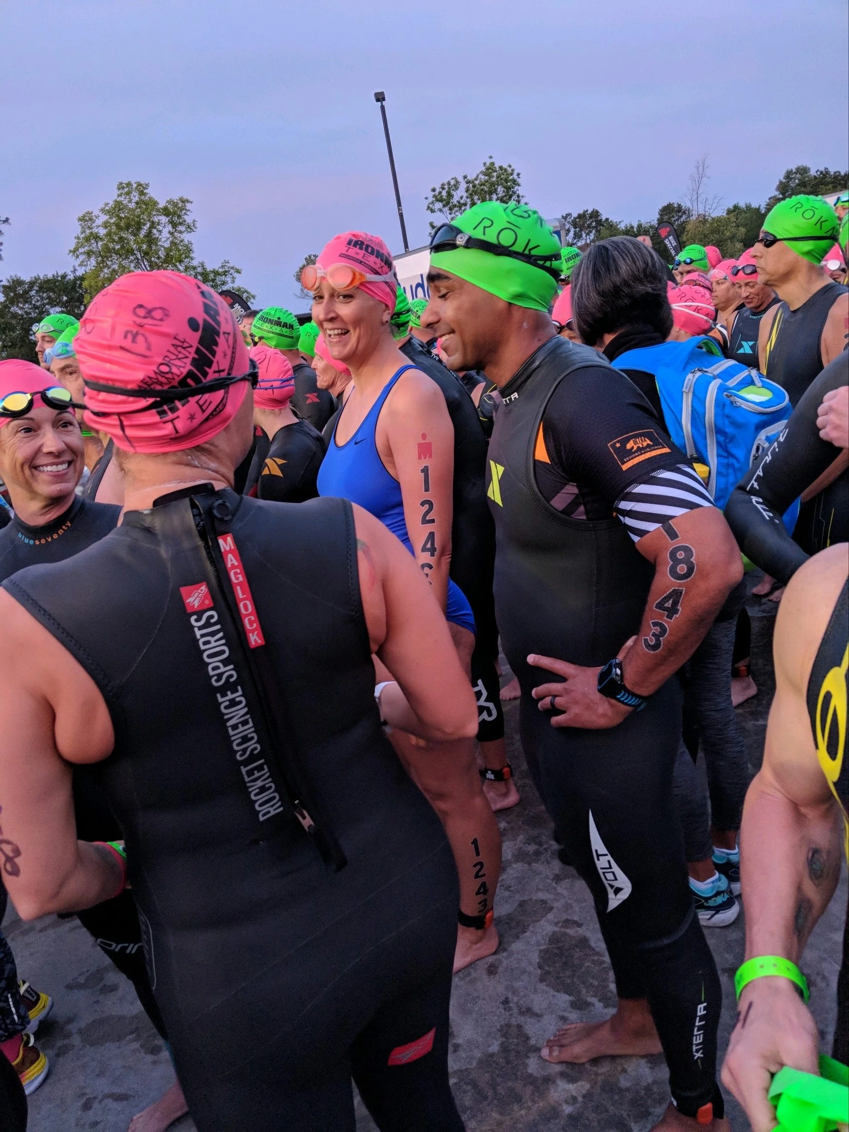 A group of athletes gathered together at the starting line of a race, wearing various sports gear including swim caps, goggles, and athletic outfits. They are smiling and interacting with each other, with some wearing neon green and pink swim caps. T