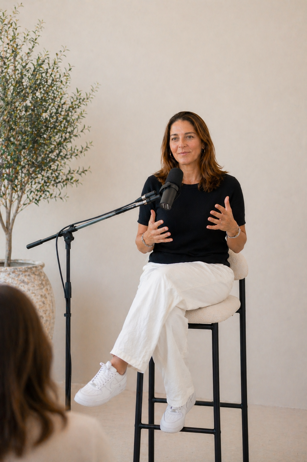 A woman with brown hair, wearing a black t-shirt and white pants, sits on a high stool and speaks into a microphone during a discussion or interview, with a potted plant beside her and an audience member in the foreground.