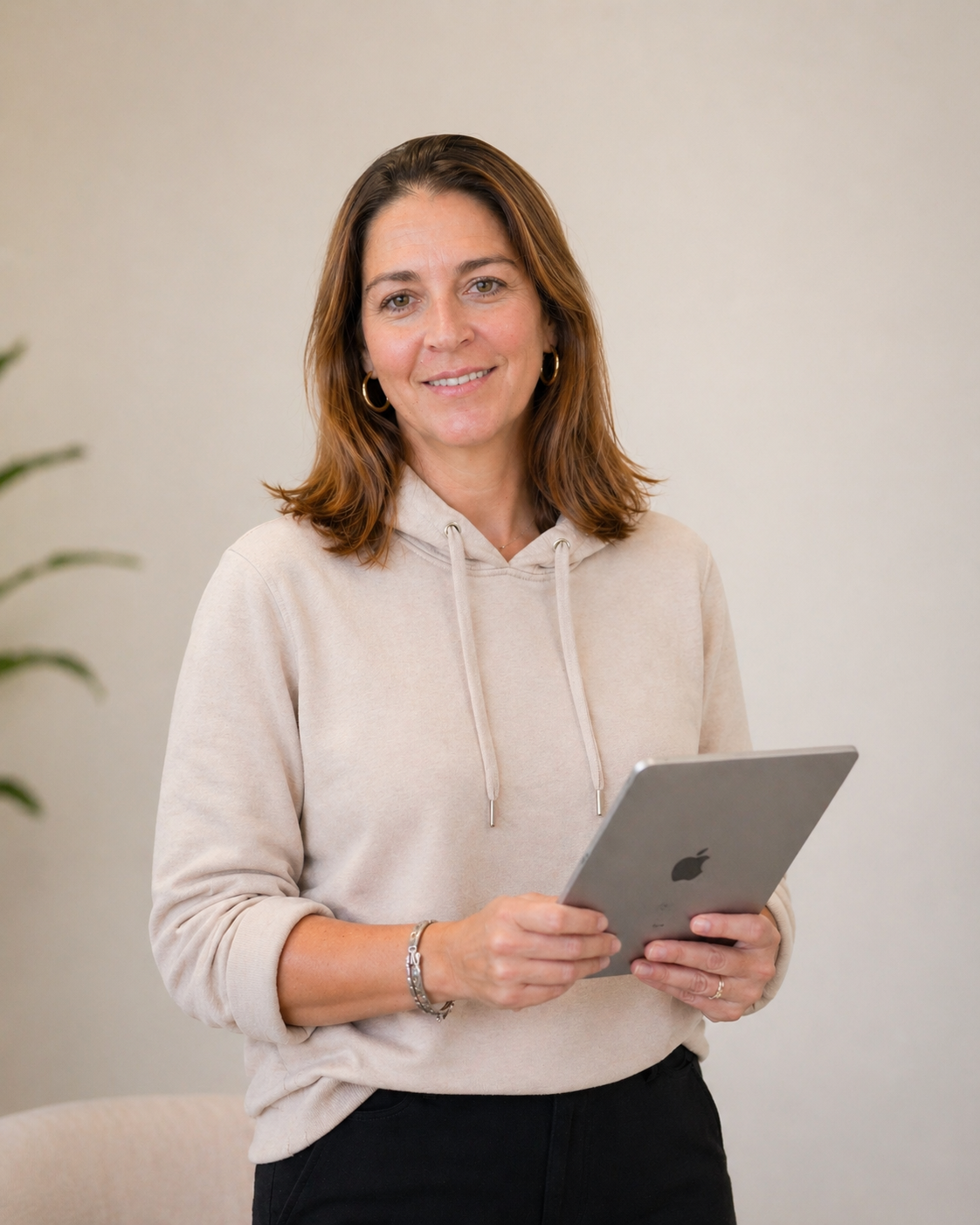 A woman with shoulder-length brown hair wearing a beige hoodie and black pants, holding an iPad, standing indoors with a neutral background.