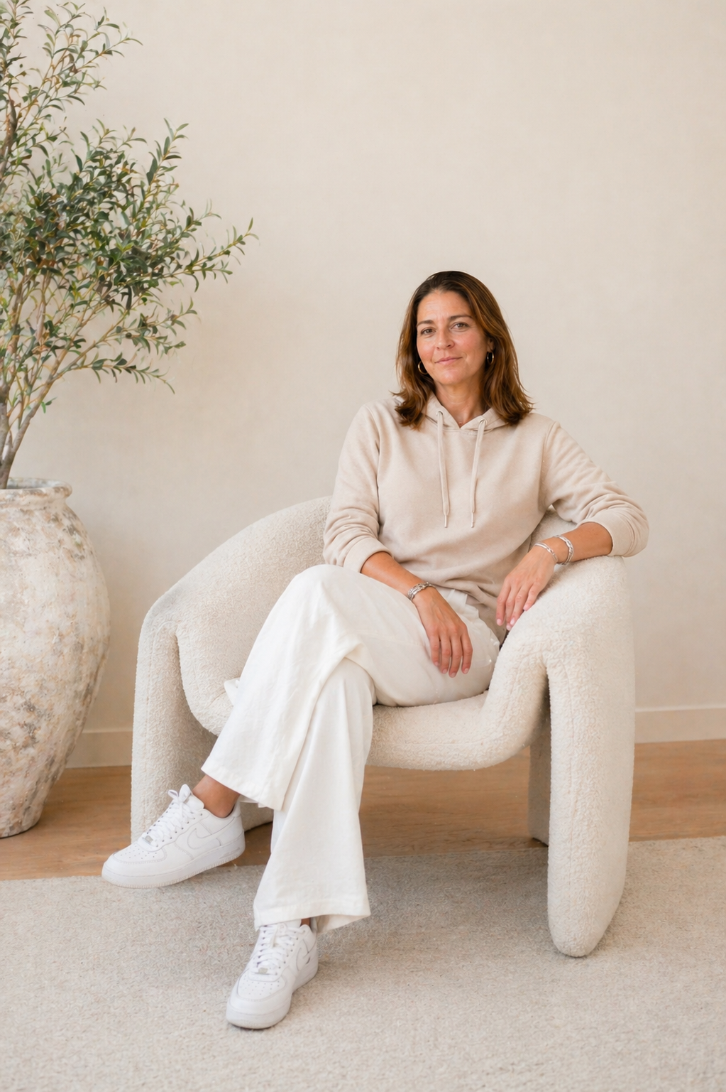 A woman sitting on a beige textured armchair in a minimalist room with a large potted plant on her left. She is dressed in a beige hoodie, white pants, and white sneakers, with a confident and relaxed expression.