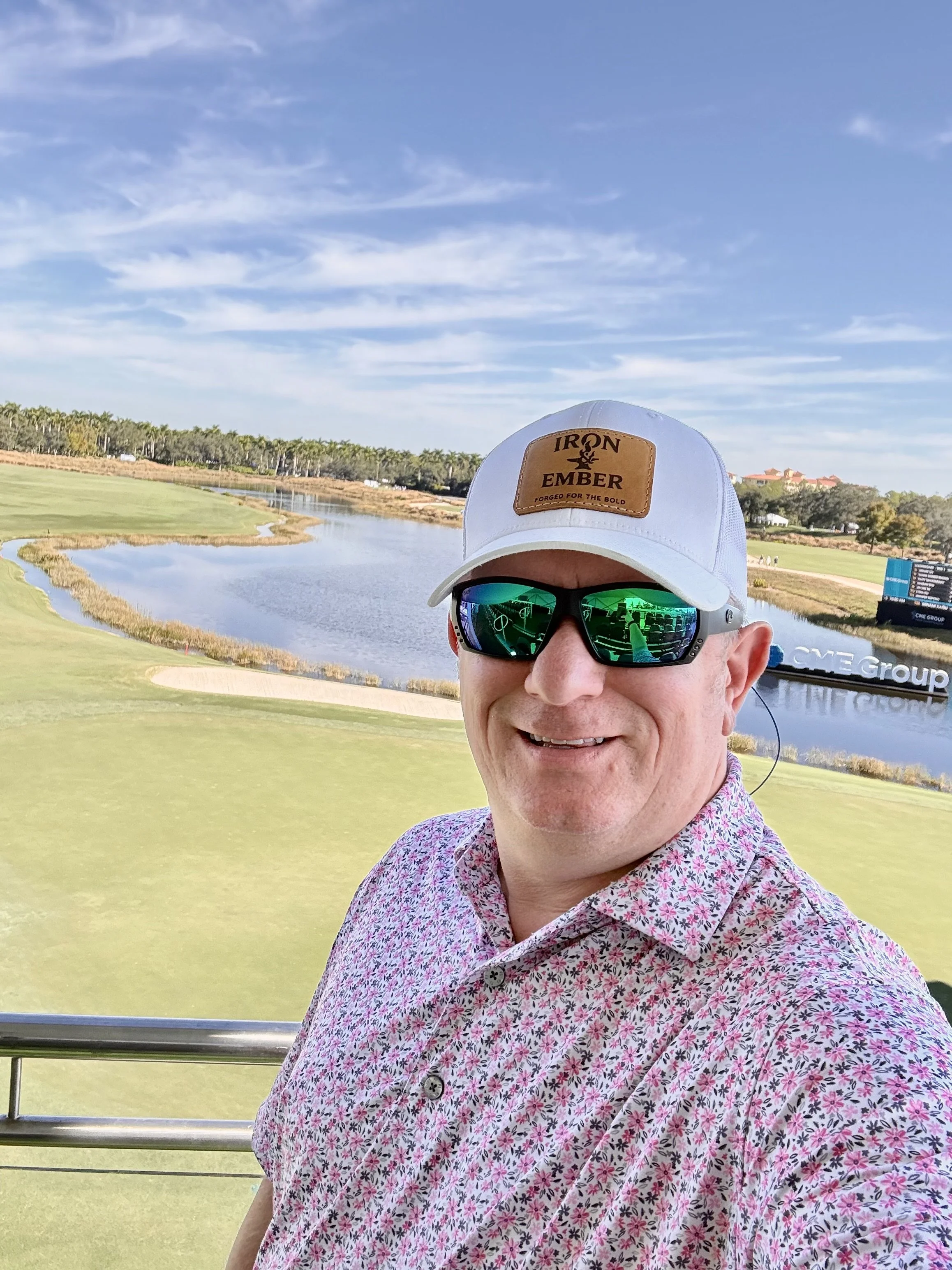 A man wearing a white cap and sunglasses with a scenic golf course and water body behind him.