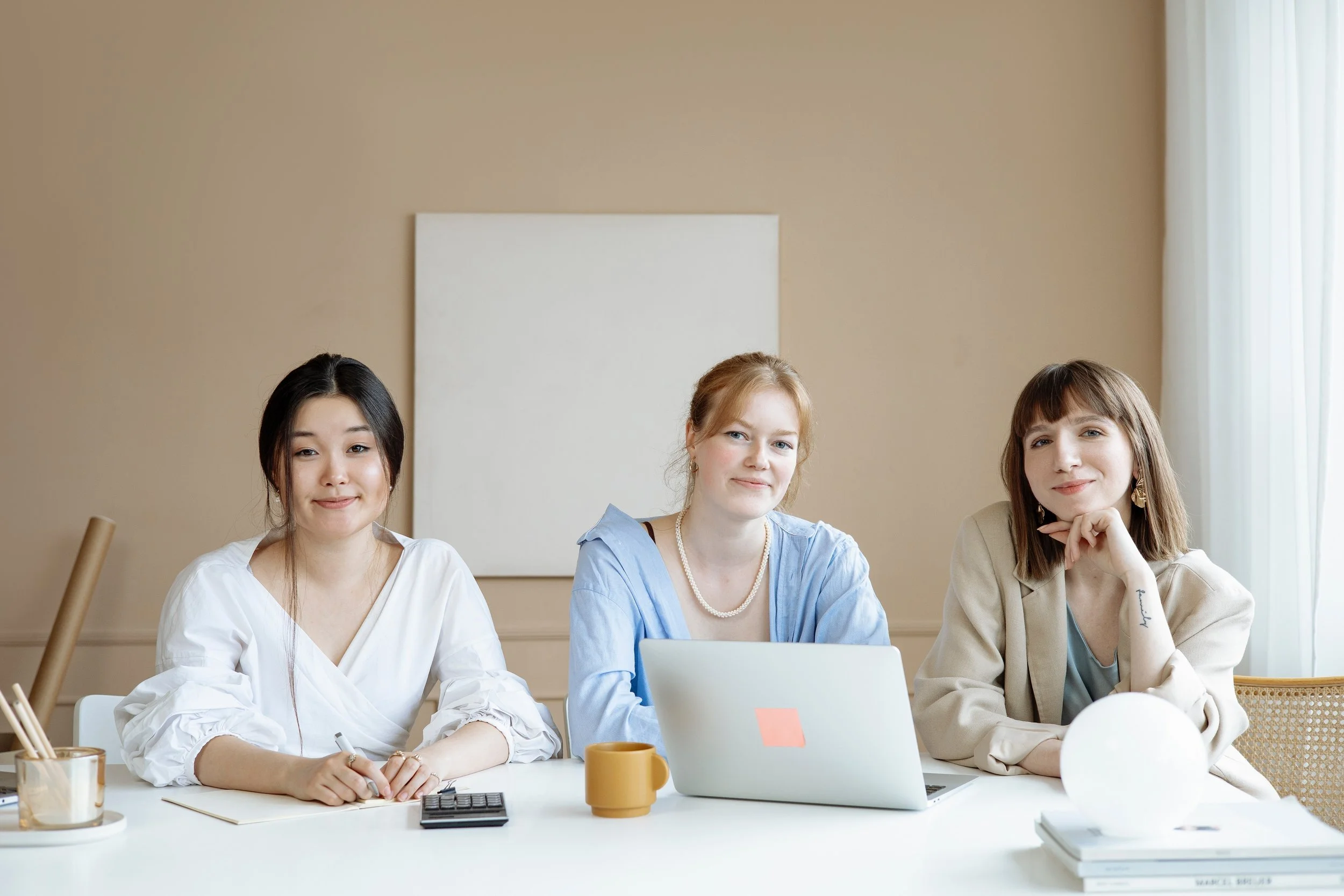 Three young women sitting at a white table in a modern room, smiling and looking at the camera. The table has a laptop, notebooks, pens, a calculator, a yellow mug, a glass, and a book. There is beige wall and white curtain in the background.