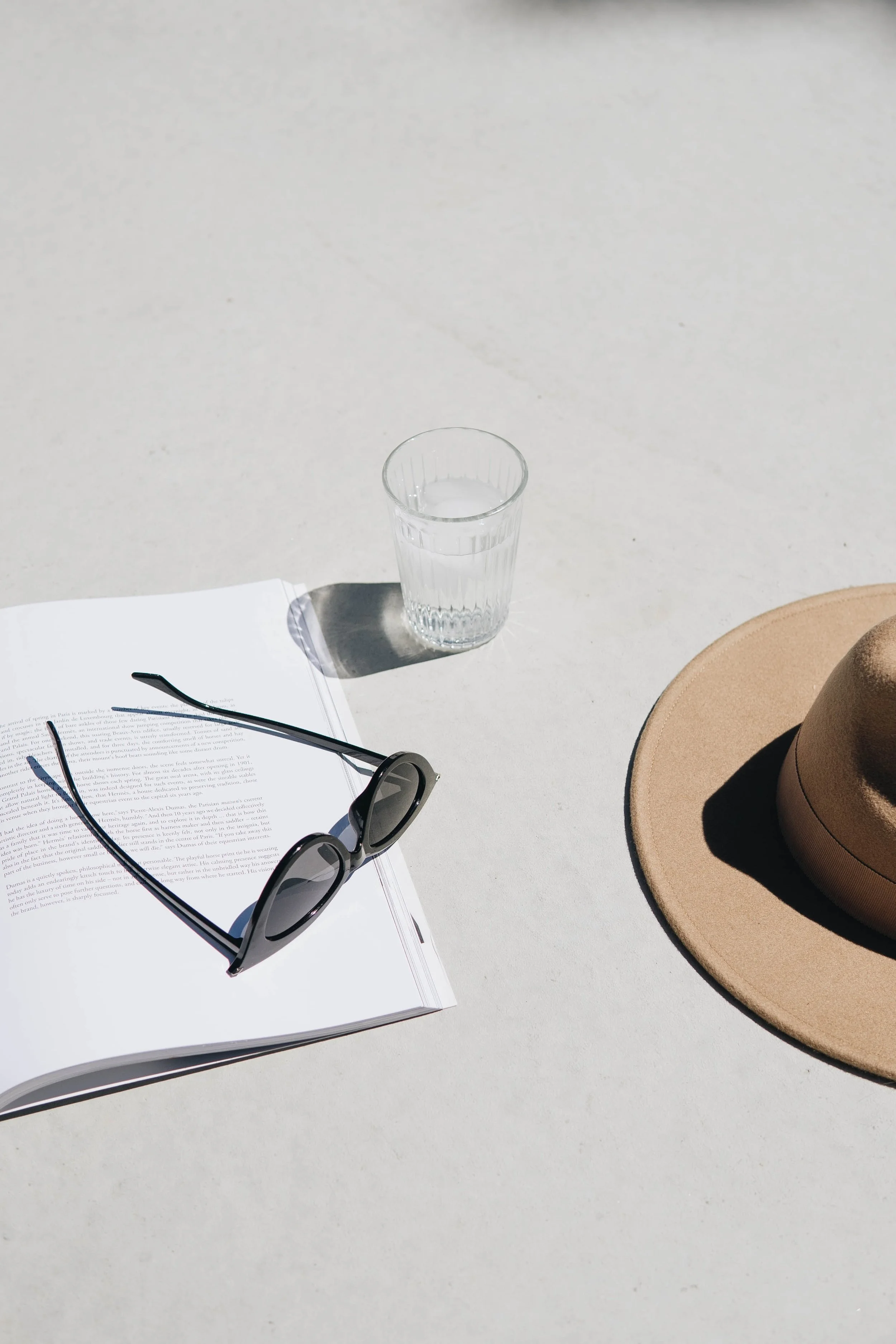 A glass of water, a pair of black sunglasses, a beige wide-brimmed hat, an open book, and a pair of black eyeglasses on a white surface, with sunlight casting shadows.