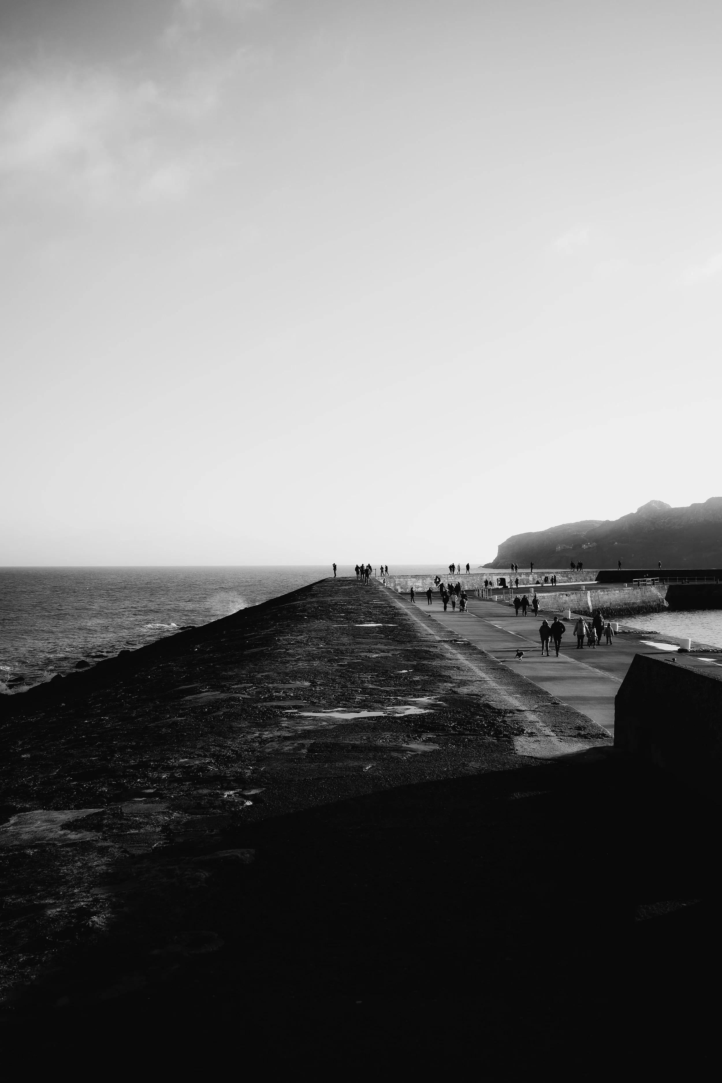A black and white photo of a pier extending into the ocean with people walking along it, a rocky hill on the right, and a cloudy sky overhead.