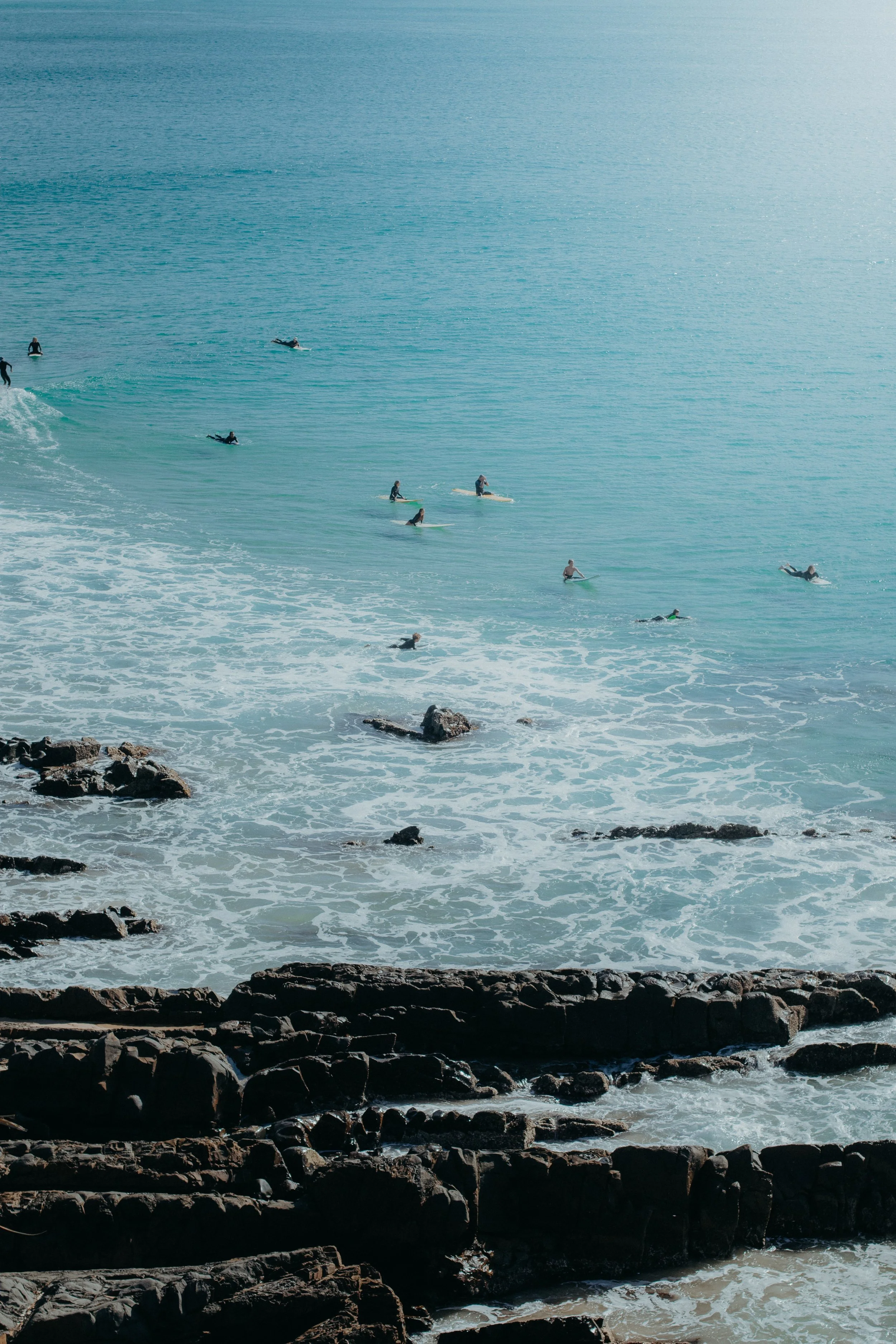 A group of people surfing and floating on surfboards in the ocean near the rocky shoreline.