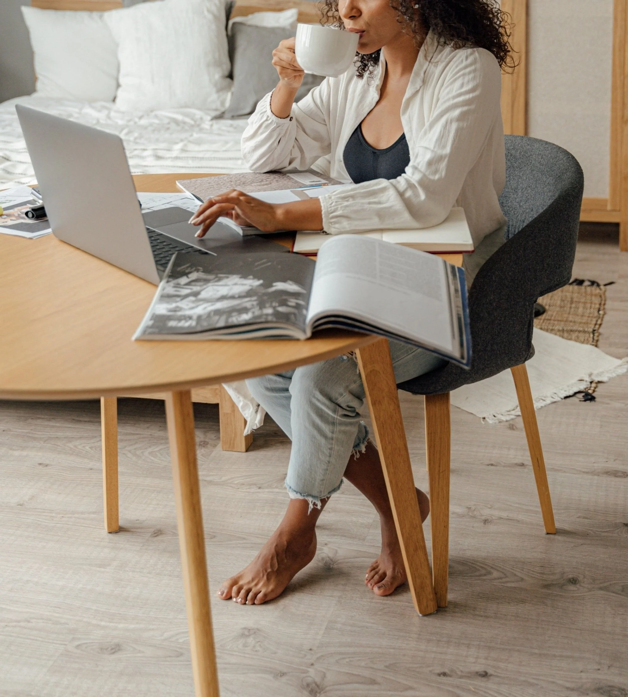 A woman with curly hair wearing a white shirt and ripped jeans is sitting at a wooden table, drinking from a large white mug. On the table, there are open magazines, papers, and a laptop. The background shows a bed with pillows and a bedside lamp.