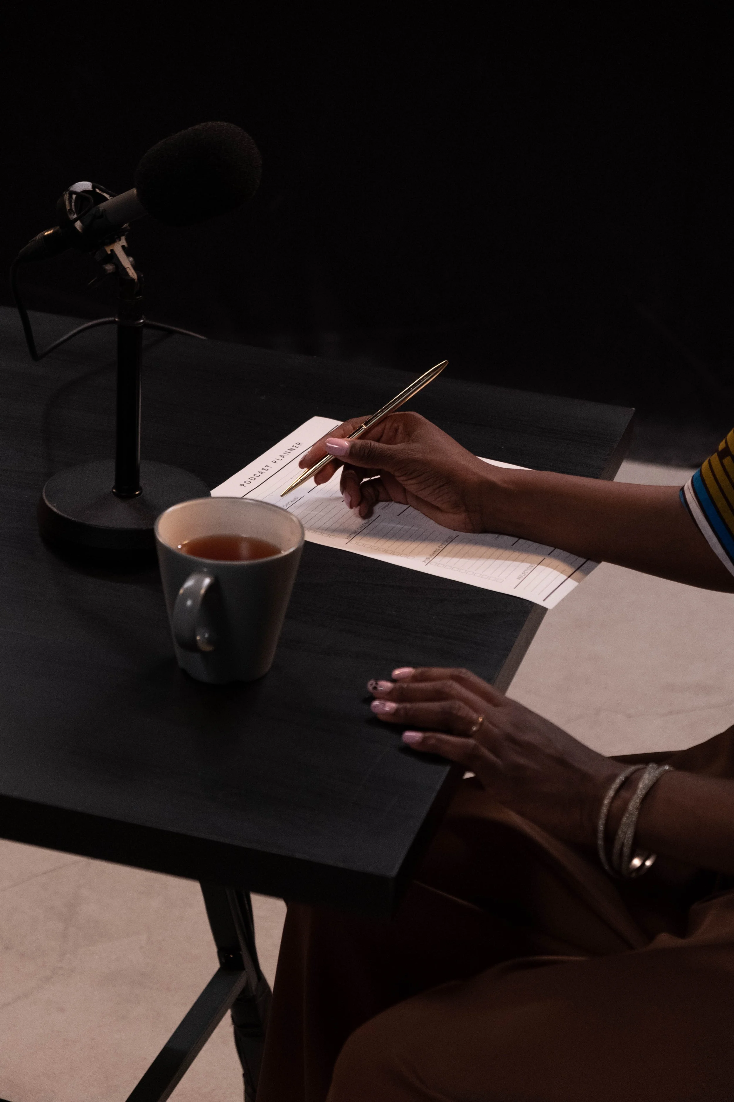 A person with dark skin and manicured nails is seated at a table, holding a gold pen and writing in a planner labeled 'PODCAST PLANNER'. On the table, there is a white ceramic mug filled with tea, and a black microphone on a stand is positioned nearby. The background is dark, and the person's arm and part of their leg are visible, with the person wearing a striped shirt and bracelet.