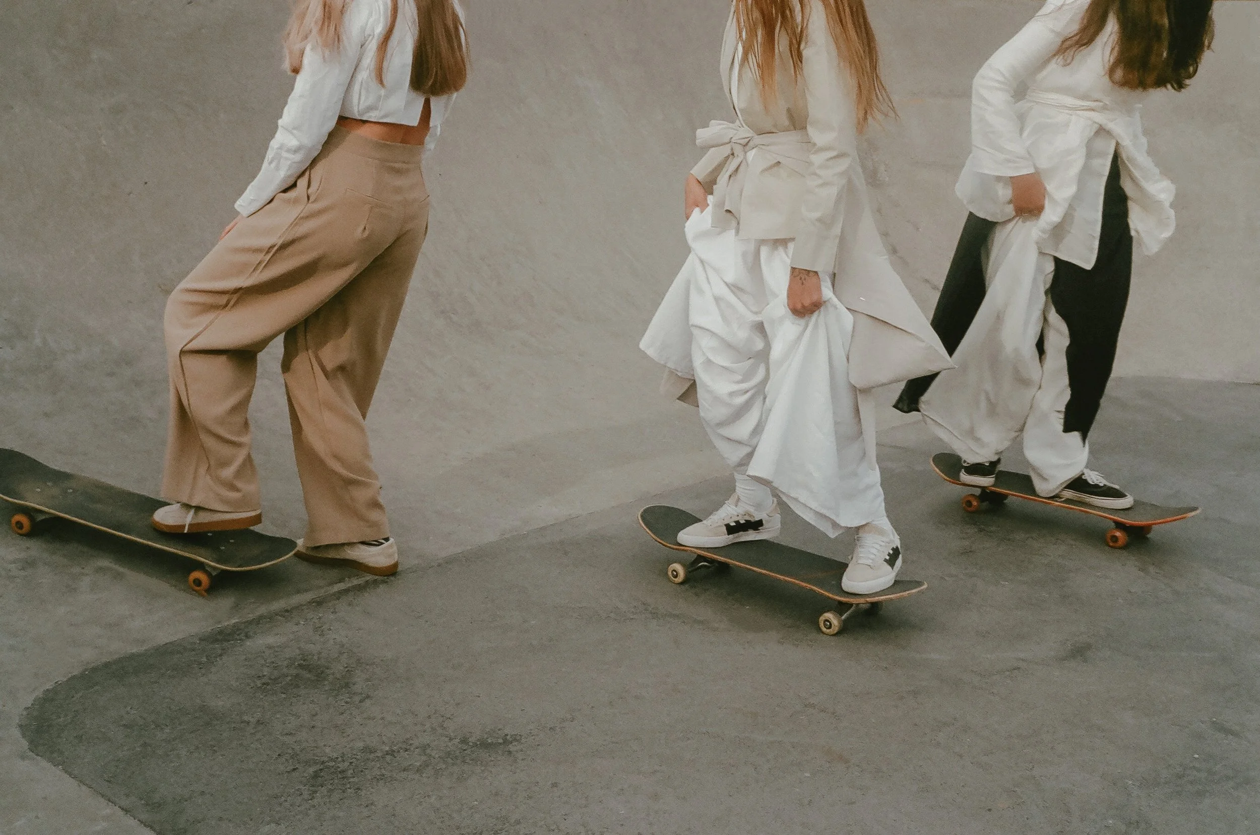 Three young women skateboarding in an indoor skate park. The first is wearing beige wide pants and a white shirt, the second is dressed in an all-white outfit with a jacket tied at the waist, and the third is in a white jacket and black pants. They are skating in a line against a plain wall.
