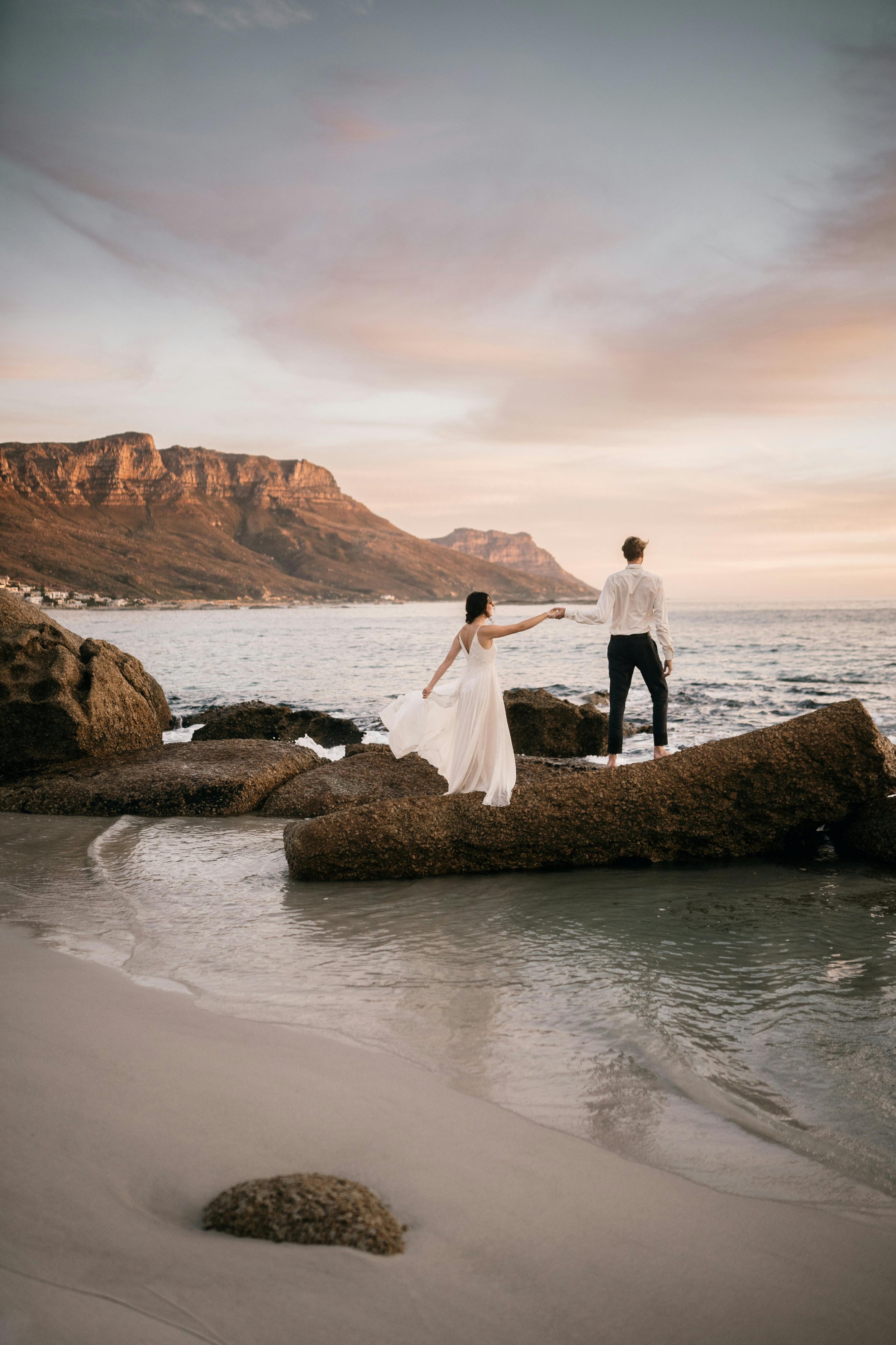 A bride and groom standing on rocks by the ocean at sunset, holding hands, with mountains in the background.