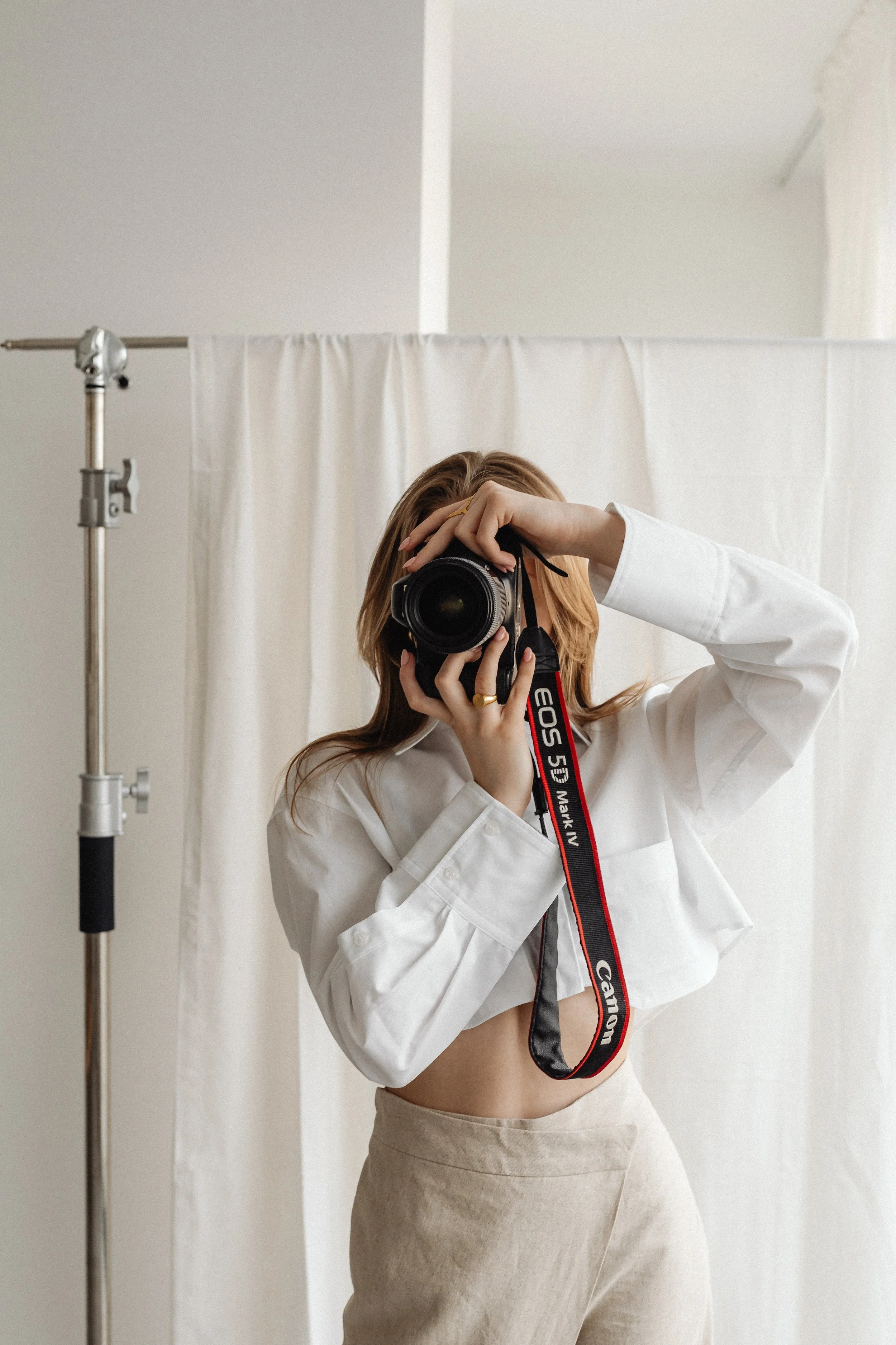 Person with long hair taking a selfie in front of a mirror, holding a camera, with a white shirt and beige pants, against a white backdrop with a curtain and a metal stand.