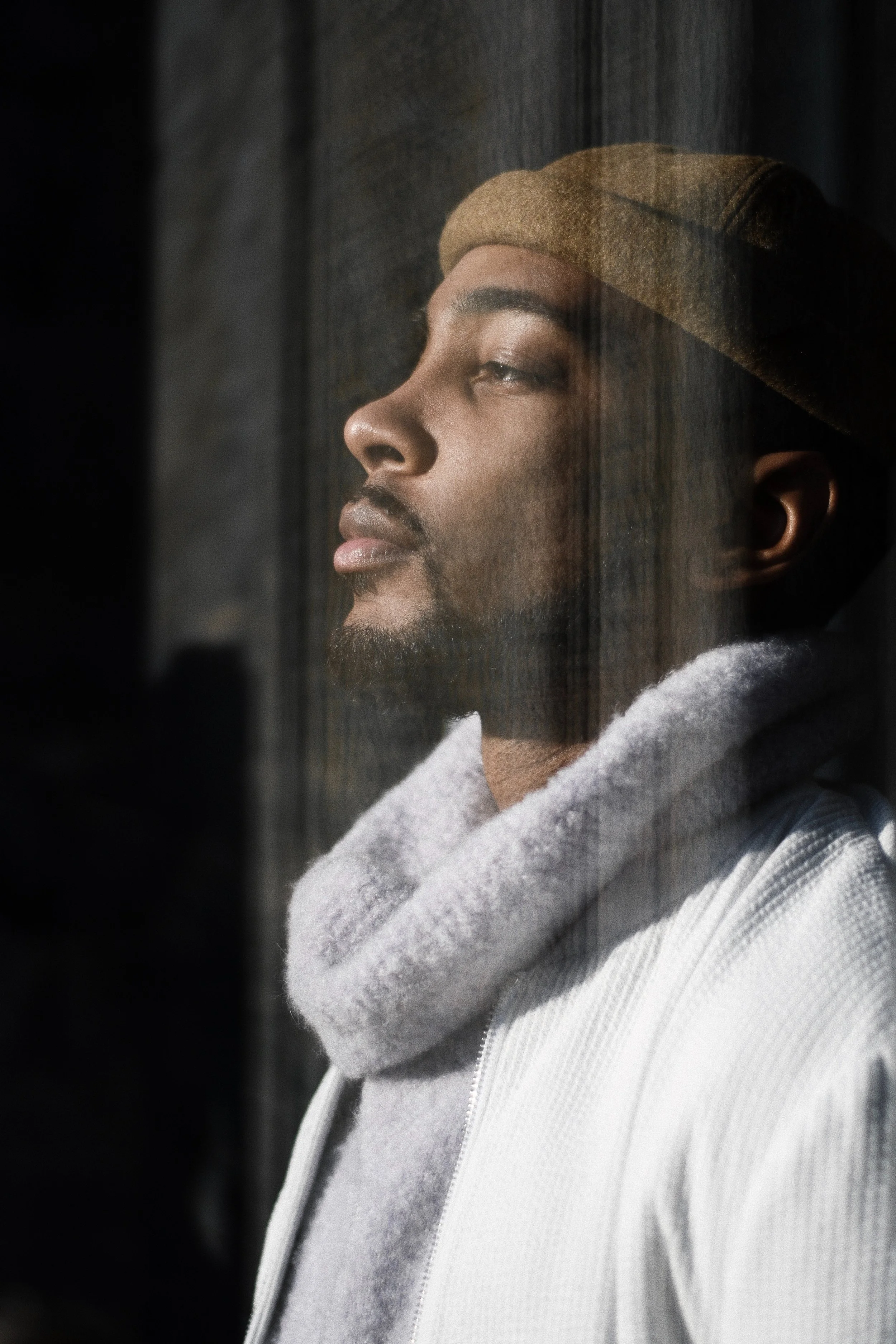 A young man with a beard and mustache wearing a beige beanie and a white hoodie, looking out a window.