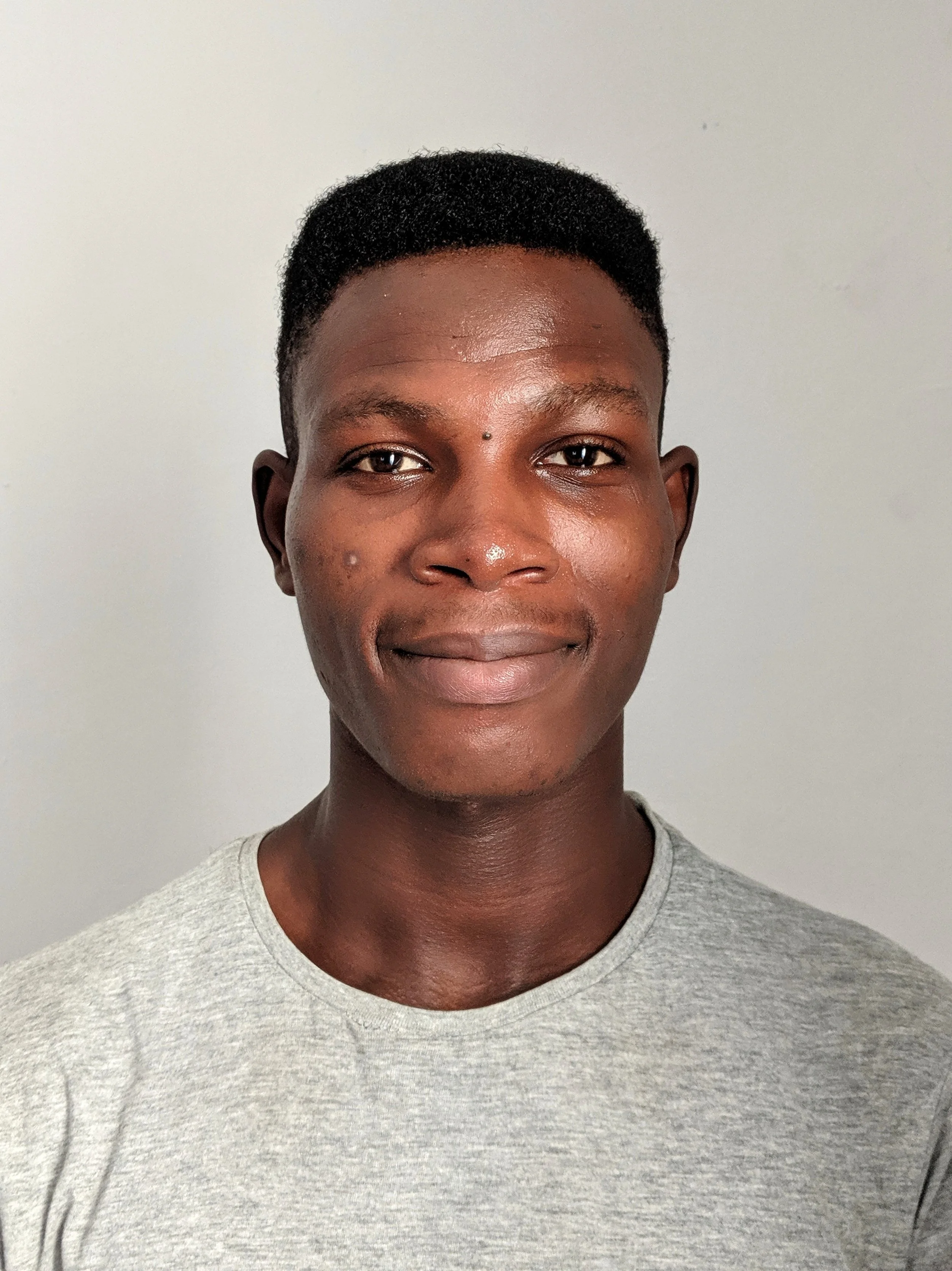 A young man with short black hair and dark skin, wearing a light gray T-shirt, standing against a plain gray background.