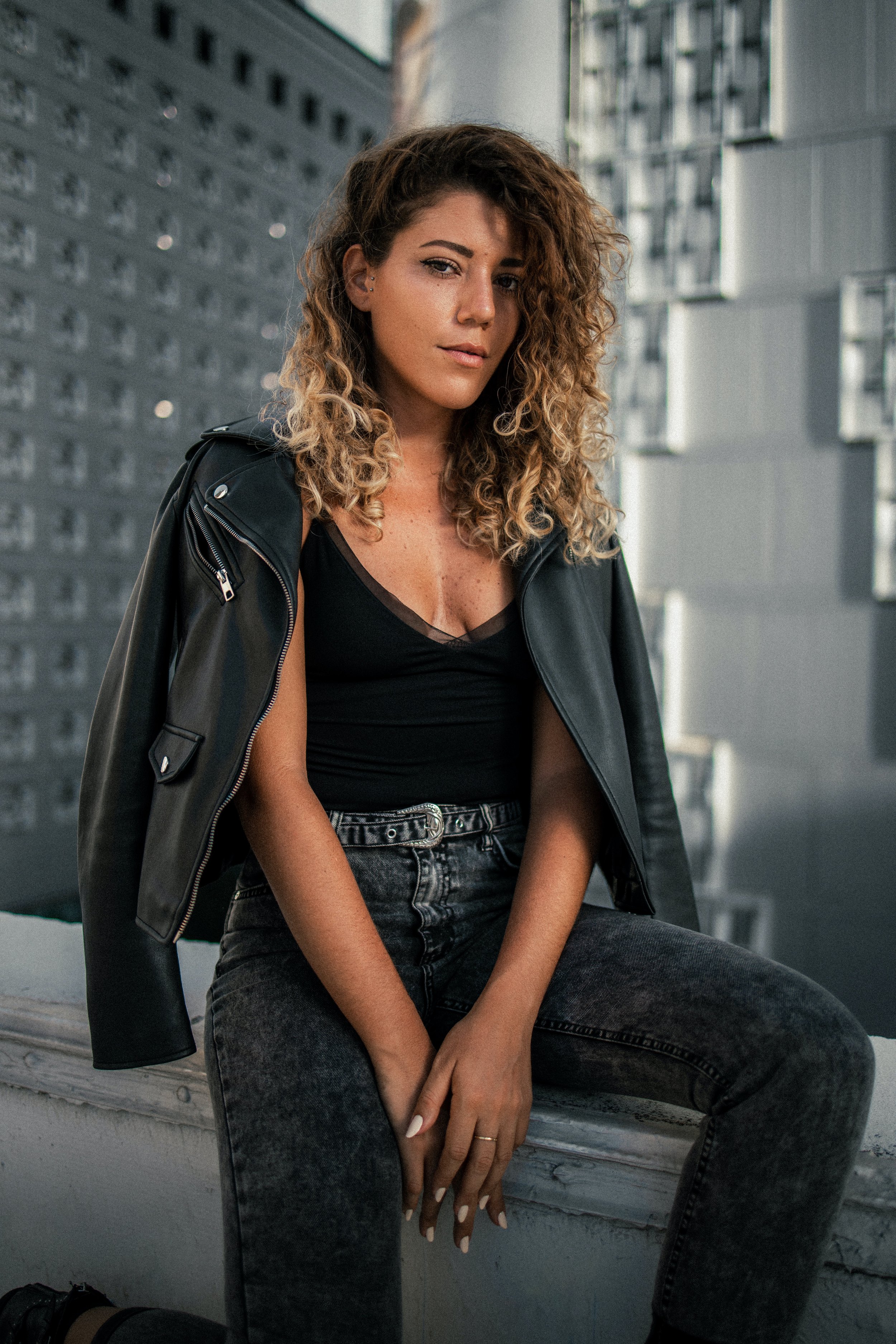 A young woman with curly brown hair sitting on a ledge outdoors with modern buildings in the background. She is wearing a black sleeveless top, high-waisted dark jeans, and a black leather jacket draped over her shoulder. She has her hand resting on her lap and is looking at the camera with a confident expression.