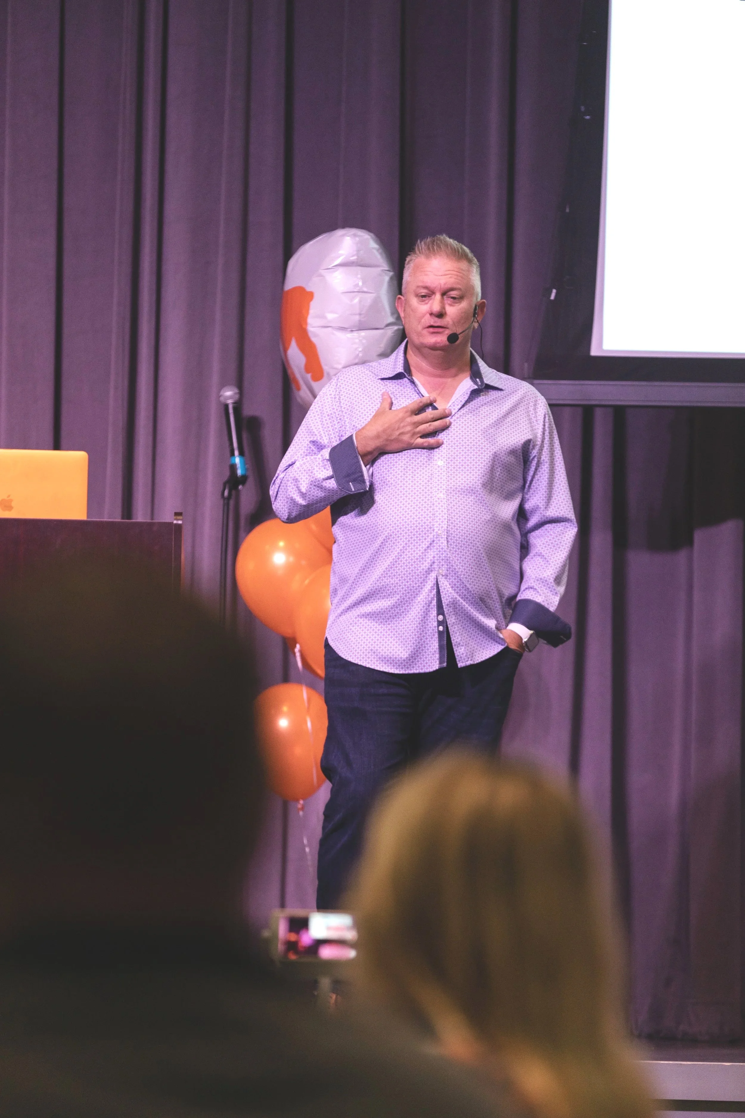 A man with short blonde hair, wearing a light-colored patterned shirt, standing on a stage with his hand on his chest. Behind him are purple curtains, orange balloons, and a large balloon with a logo. A screen displaying white space is visible in the