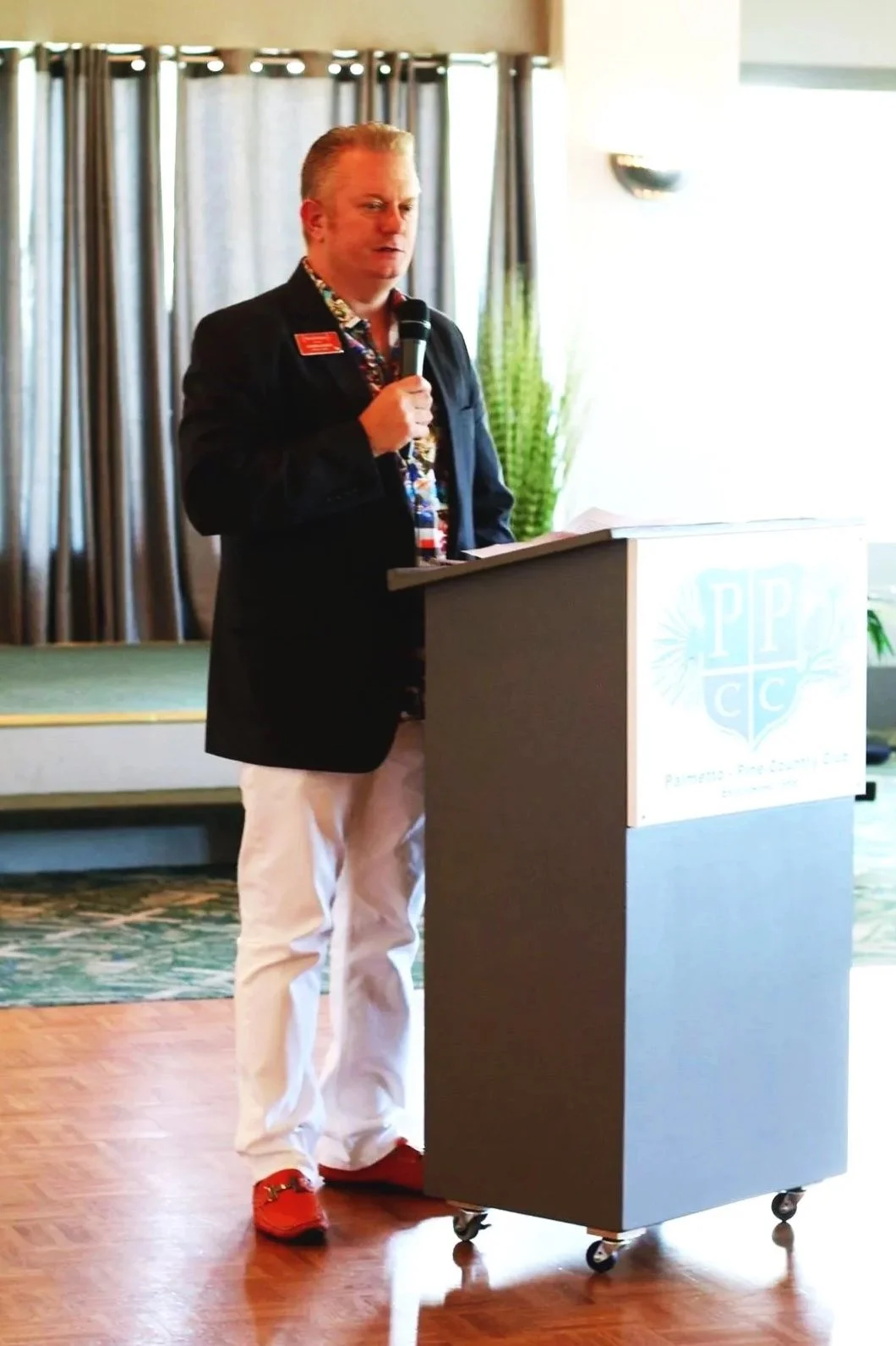 A man with a microphone standing behind a podium, delivering a speech at an indoor event. He is wearing a black blazer, a colorful shirt, white pants, and red shoes. Behind him are curtains and some plants.