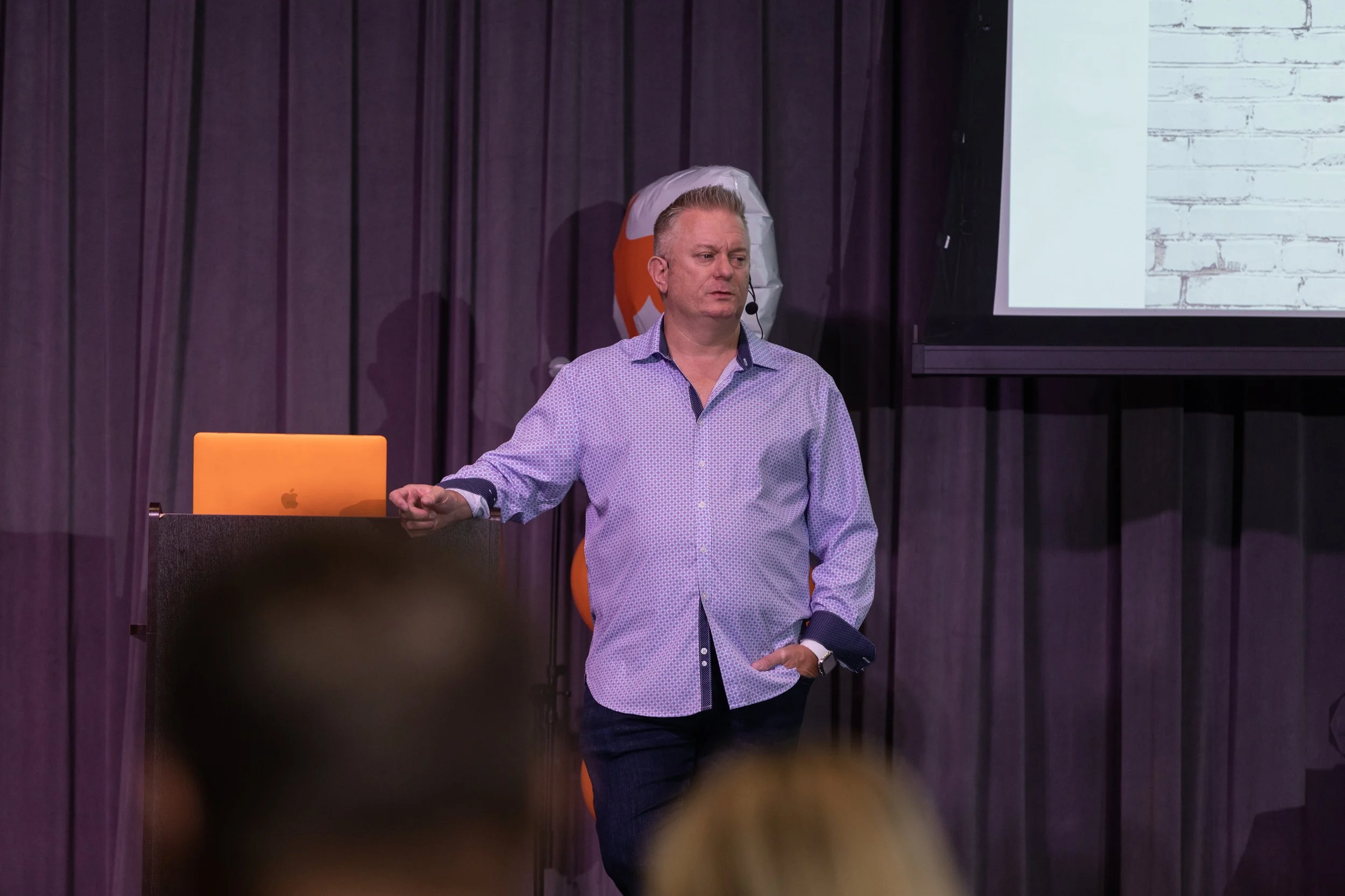 Jason Jakus National Speaker in a light blue button-up shirt and dark jeans standing while giving a presentation in front of a purple curtain and a screen with a whiteboard background.