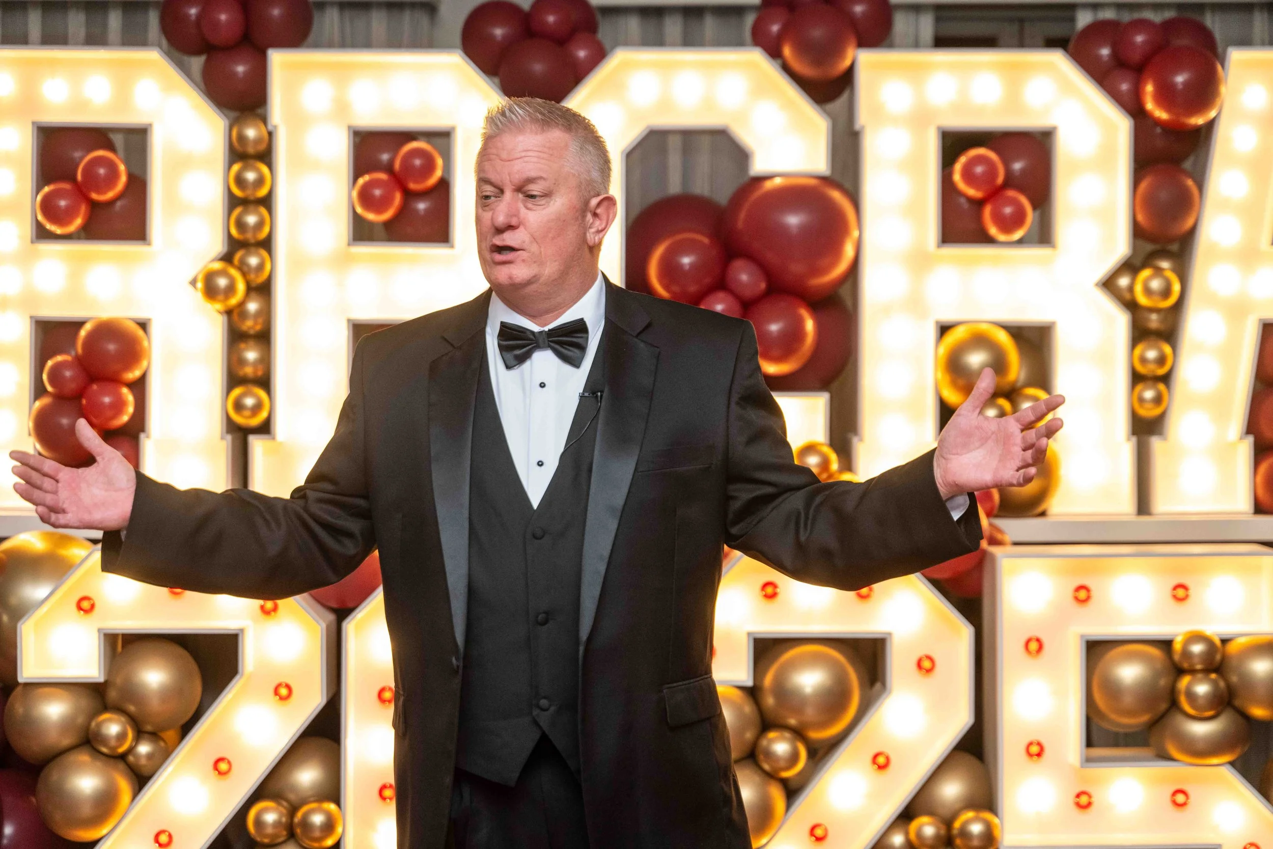 A man in a tuxedo speaking with his arms outstretched in front of a decorative backing with large illuminated numbers and clusters of red and gold balloons.