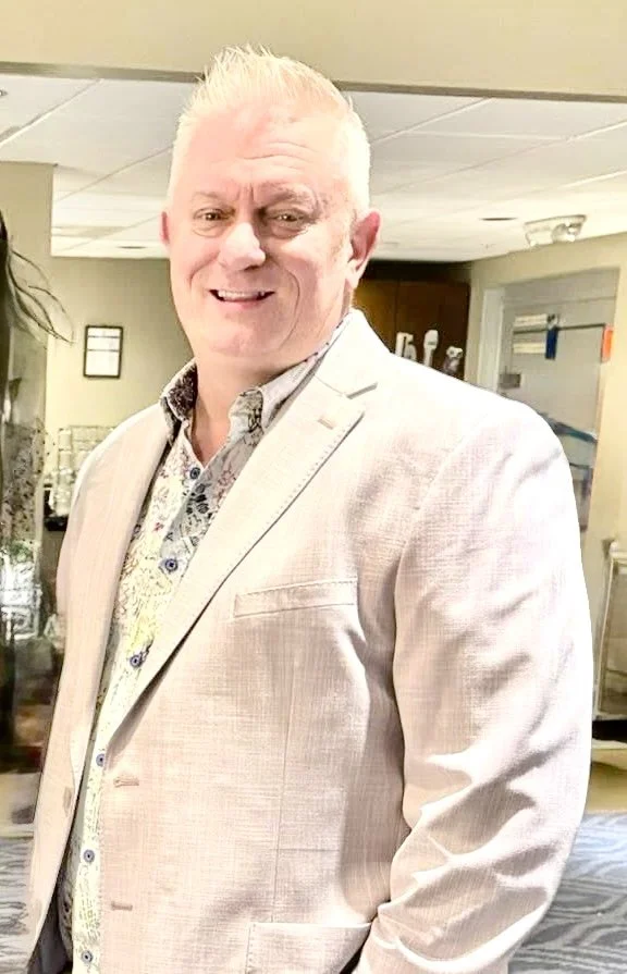 Smiling man with white hair wearing a light-colored blazer and patterned shirt in an indoor setting.