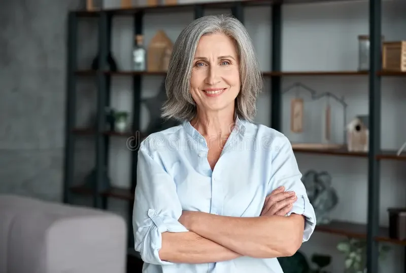 An older woman with gray hair smiling and crossing her arms in a modern living room or office with shelves in the background.