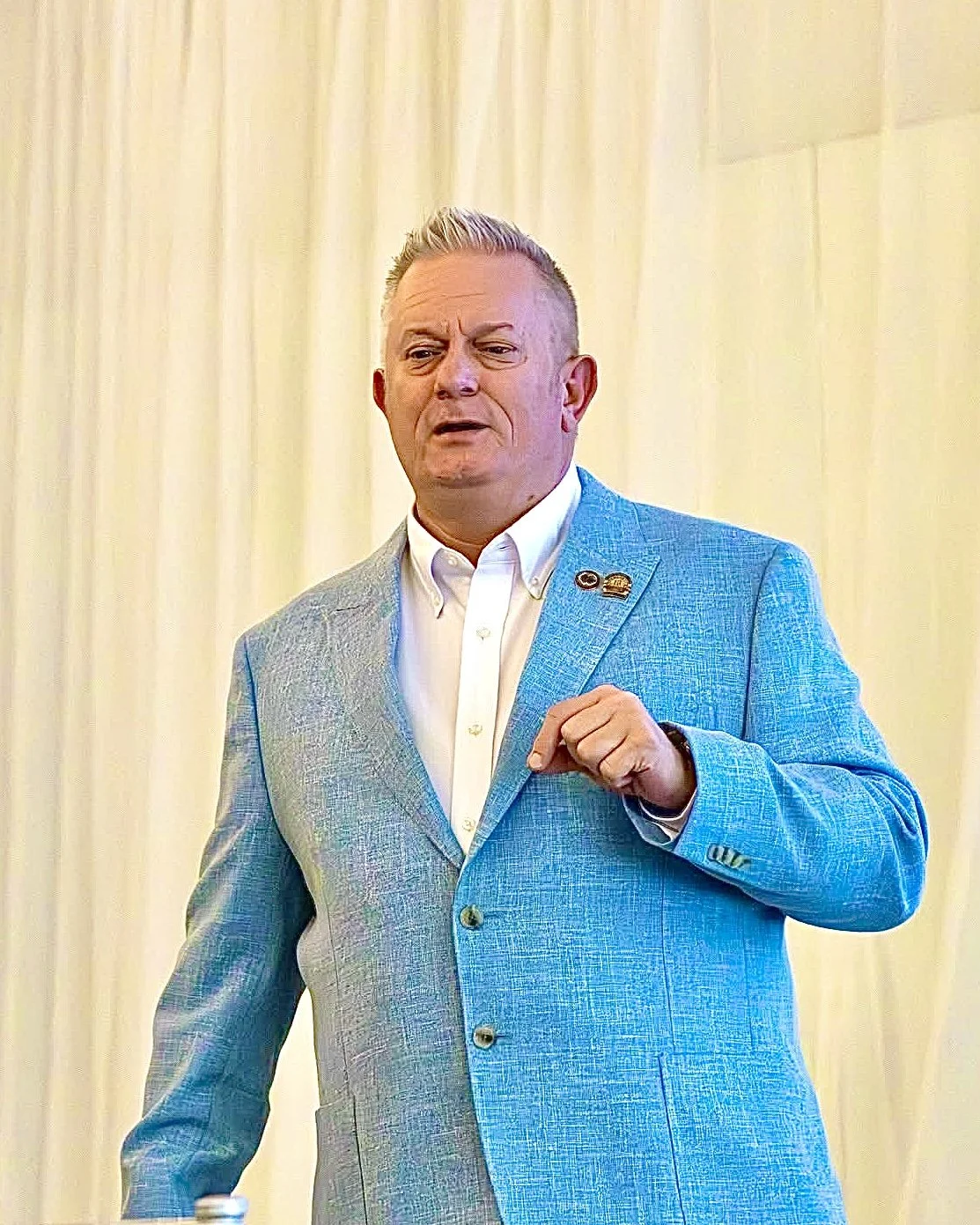 Jason Jakus National Speaker in a blue suit jacket and white shirt speaking or gesturing during a presentation or speech in front of a cream-colored curtain.