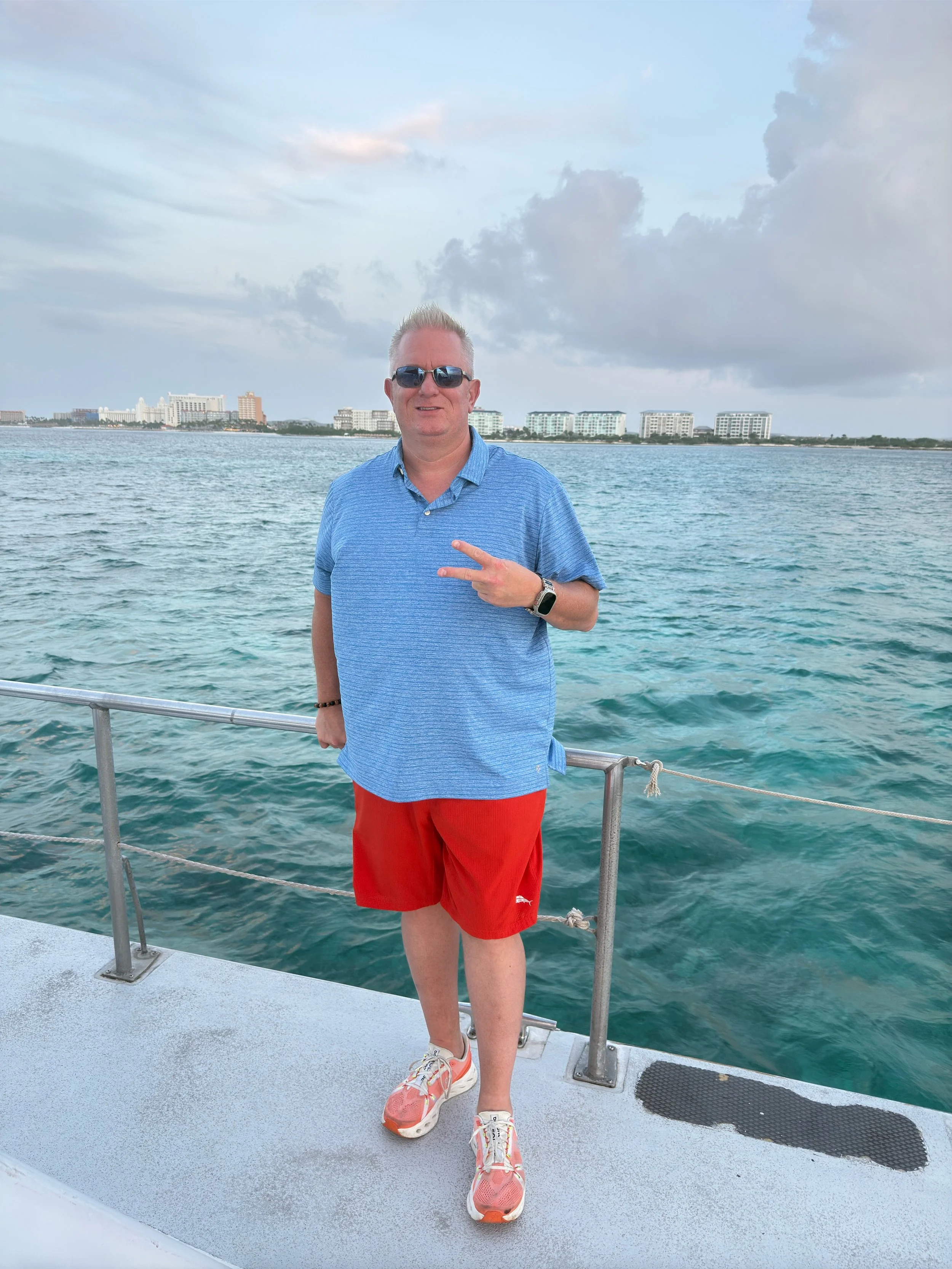 Jason Jakus wearing sunglasses, a blue shirt, red shorts, and sneakers standing on a boat with water and buildings in the background, making a peace sign.